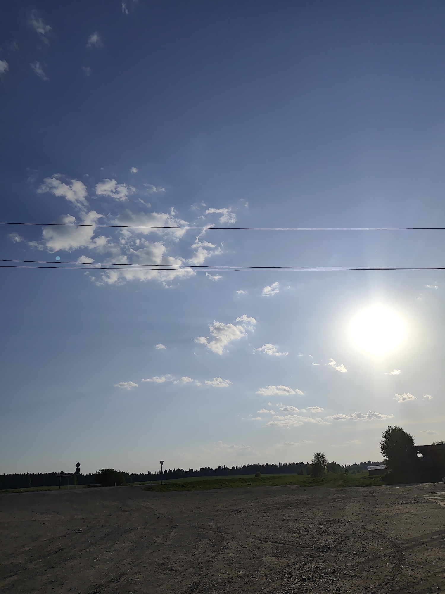 The image shows a rural scene near Нижний Пальник, Russia. The foreground is a dirt road or open area with textured, gravelly ground. Beyond the road, there's a green field with sparse vegetation. In the middle ground, a line of trees and bushes forms a distant treeline against the sky. Two road signs are also visible in the center. The sky takes up the majority of the frame, with scattered white clouds and power lines. The sun is bright on the right side, shining through some thin clouds, creating rays of light.