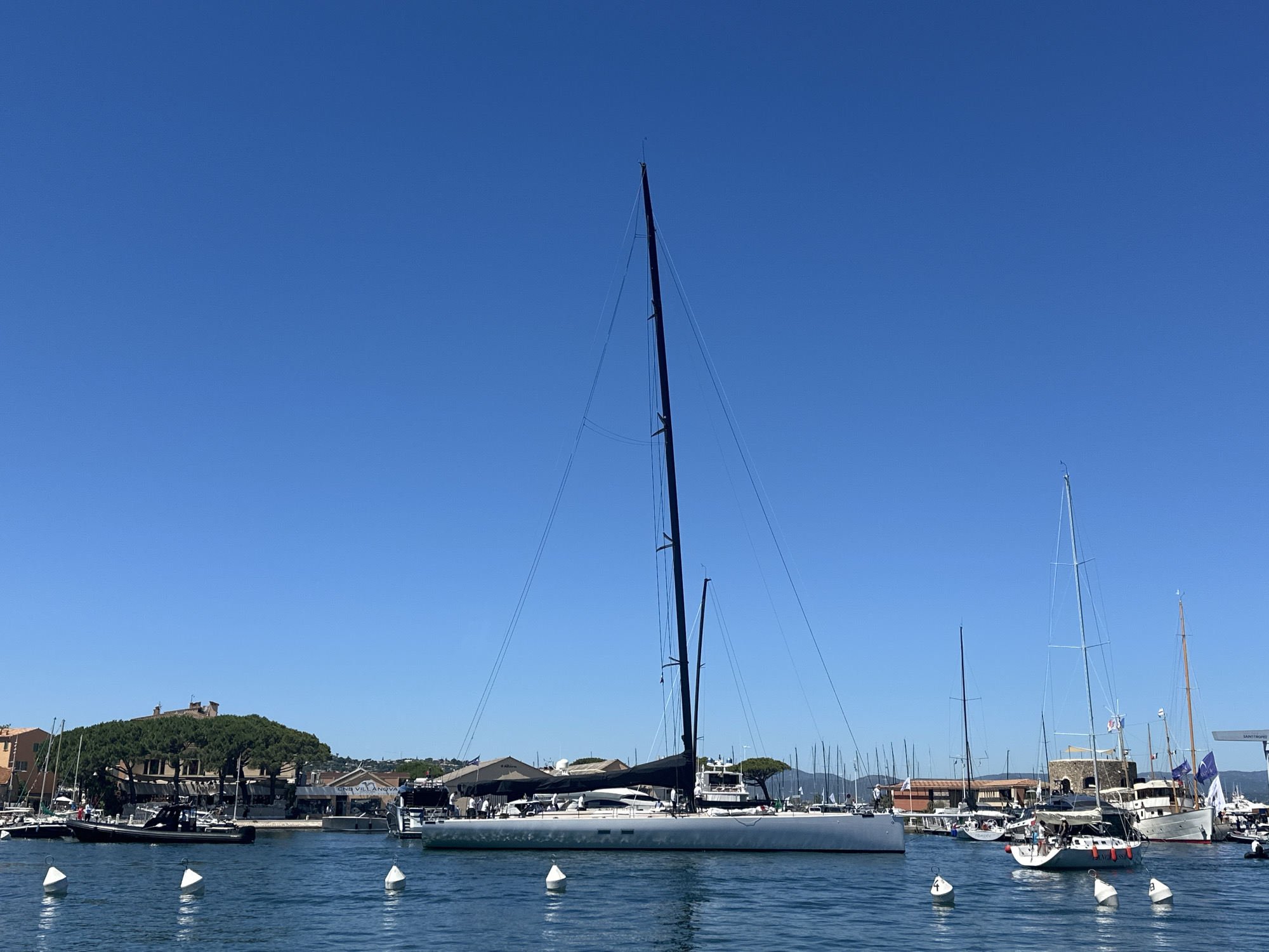 The image captures a sunny day in the port of Saint-Tropez, France, with a clear blue sky dominating the upper portion of the frame. In the foreground, the calm blue water of the harbor is dotted with several white buoys.
The central and most prominent subject is a large, modern sailboat with a sleek, light-colored hull and a tall, dark mast. Its black mainsail is furled. The boat appears to be moored alongside other vessels.
To the left of the large sailboat, a black inflatable boat with a white stripe is navigating through the water, carrying several people. Further in the background, a cluster of buildings with terracotta roofs are visible, interspersed with lush green trees. One building, seemingly a historic structure, stands out with its architecture.
To the right of the large sailboat, numerous other sailboats of varying sizes are docked. Some have their sails down, while others have their masts clearly visible against the sky. A larger, white yacht with multiple decks and flags is also present in this section of the harbor. The overall scene depicts a bustling yet serene maritime environment.