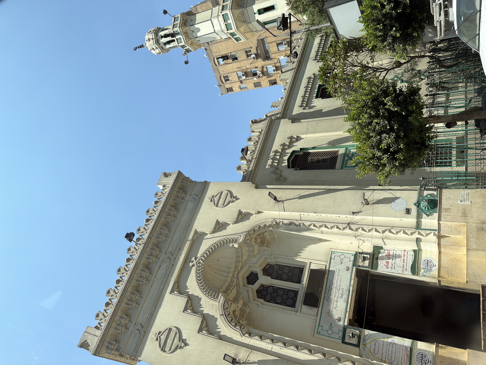 The image shows the exterior of the Mosque of al-Aqmar, located in Islamic Cairo, Egypt. The mosque, built in the Fatimid architectural style, features a unique facade with intricate geometric carvings and muqarnas. The main entrance is visible with Arabic inscriptions and banners. To the right, a minaret of a nearby mosque, likely the Mosque of al-Ghuri, can be seen against a clear blue sky. In the background, a modern apartment building stands, contrasting with the historical architecture. Trees and greenery line the street in front of the mosque, and a white car is partially visible in the foreground. The scene is bathed in bright sunlight, indicating a daytime setting.