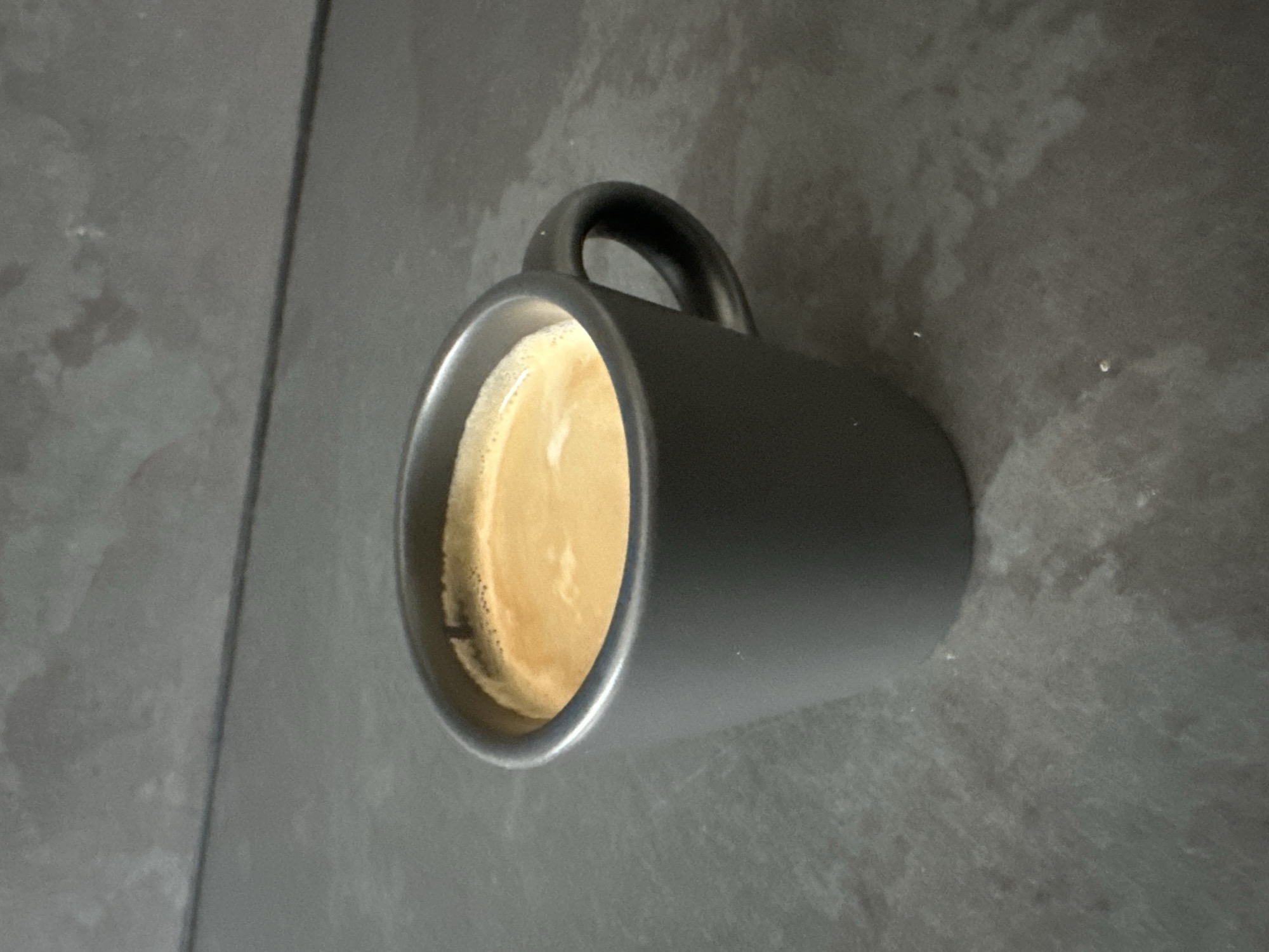 A black mug of coffee sits on a dark, textured surface. The coffee has a creamy, light brown crema on top, with some darker specks visible at the edge of the mug. The mug has a simple, curved handle. The background is a mottled gray, possibly a countertop or table, with a slight sheen. A thin, dark line runs diagonally across the left side of the frame, suggesting a seam or the edge of another surface. The lighting is soft, and the overall impression is one of a quiet, possibly morning, moment. There is no visible text in the image. The location context is Belfast City, United Kingdom.