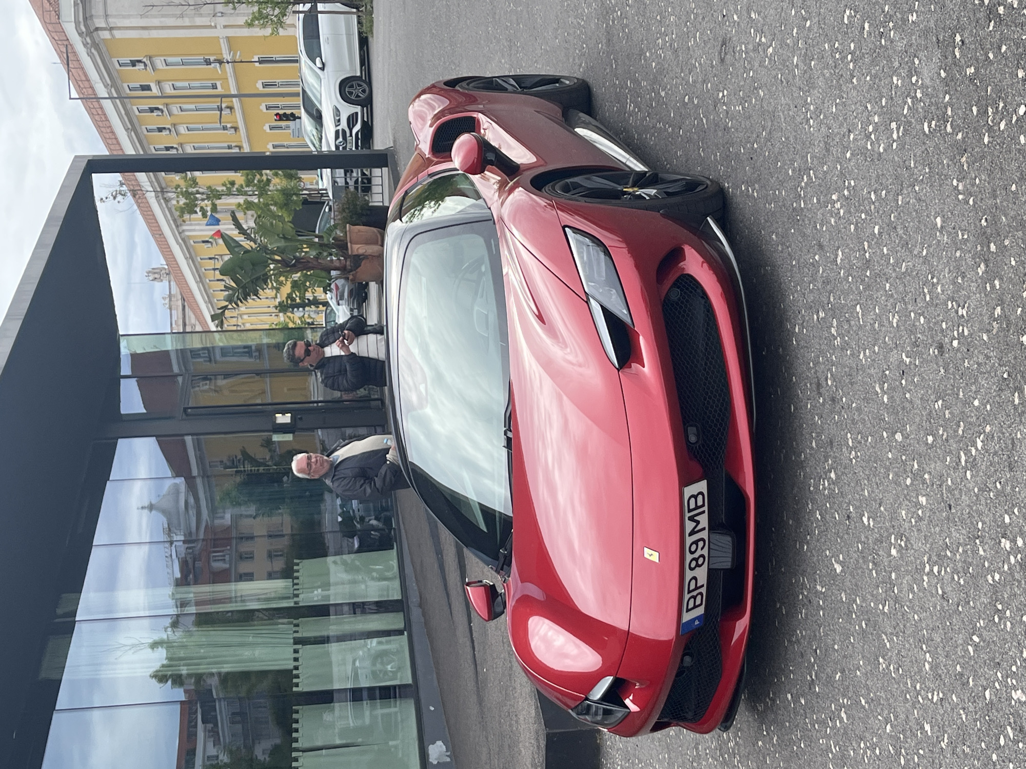 A red Ferrari sports car is parked on the side of a street in Lisbon, Portugal. The license plate reads "BP 89 MB." An older man in a dark jacket with a white collar stands near the car, reflected in the glass windows of a modern building next to the vehicle. A second person in a dark jacket is visible in the reflection behind the man. A yellow building is across the street, partially obscured by foliage. A white car is parked further down the street. The sky appears overcast.