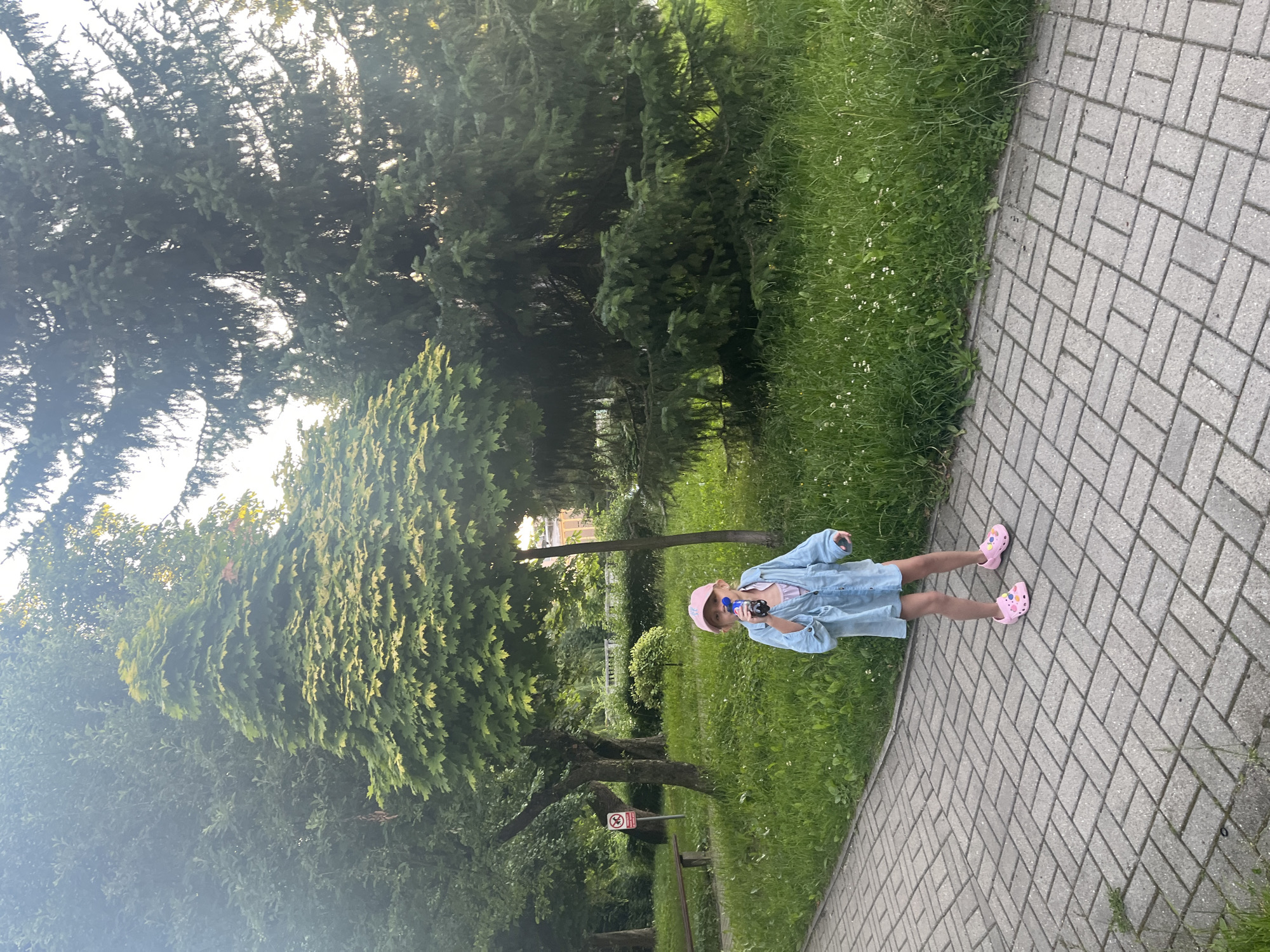 A young girl wearing a pink cap, a light blue shirt, and pink clogs stands on a paved walkway in a park. She is holding a dark bottle in her right hand and appears to be drinking from it. The girl is positioned in front of a grassy area with several trees and a small, decorative bush. To the left of the girl, a wooden bench and a circular sign with a red border and a white diagonal line are visible. In the background, larger trees, including a tall evergreen, fill the scene. The lighting suggests it is late afternoon or early evening.
