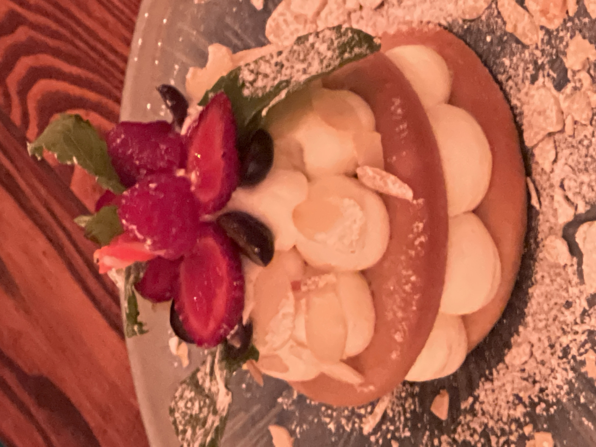 A close-up shot captures a decadent dessert, possibly a mille-feuille or layered cake, resting on a dark blue plate. The dessert is composed of two circular, golden-brown layers, possibly biscuits or thin cakes, sandwiching generous dollops of creamy white filling. The filling is artfully piped and topped with fresh red raspberries and sliced strawberries, with a few dark blueberries interspersed. Delicate green mint leaves provide a vibrant garnish, and slivers of almonds are scattered amongst the cream. A dusting of powdered sugar lightly coats the entire confection and the surrounding plate, with some larger crumbs scattered on the plate as well. The background features a warm, reddish-brown textured surface, likely a wooden table, adding to the cozy and inviting atmosphere. The lighting is warm and focused on the dessert, creating a slight bokeh effect on the background. The image does not contain any individuals or specific landmarks that would indicate its location in Novosibirsk, Russia.