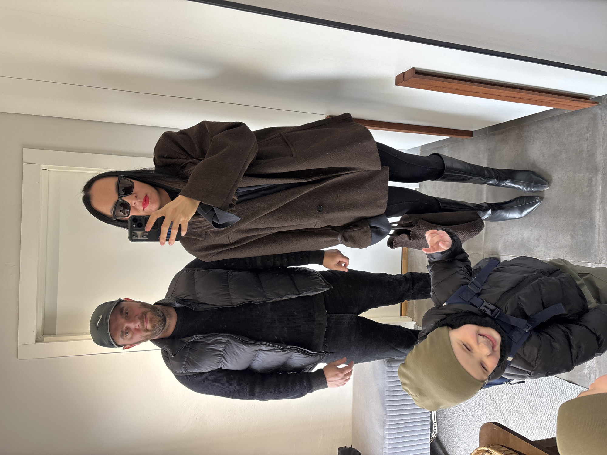 A woman, a man, and a young child are posing for a selfie in front of a mirror, likely in a retail setting. The woman, positioned in the upper part of the image, is wearing sunglasses, a brown coat, leggings and black boots, and holding a phone. The man, slightly below her, is wearing a black vest, a sweater and a hat. The child, at the bottom of the image, is smiling and wearing a hat and a puffy jacket with a backpack. The background shows portions of the retail space, including the door and some furniture. There is no indication in the image that can identify Chaikovsky, Russia as the location.