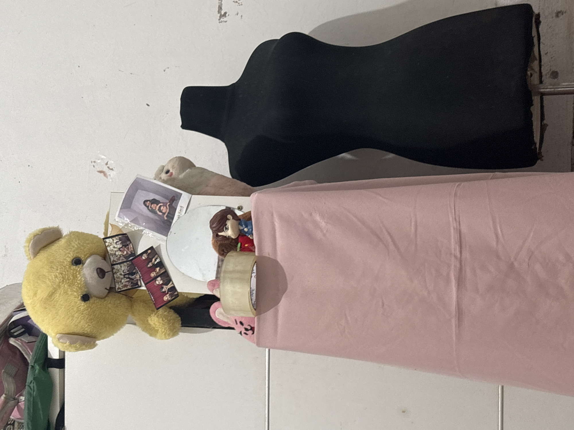 A stuffed yellow teddy bear is holding a collection of photos and trinkets. It is placed on top of a pink draped table. Next to the table is a black mannequin.