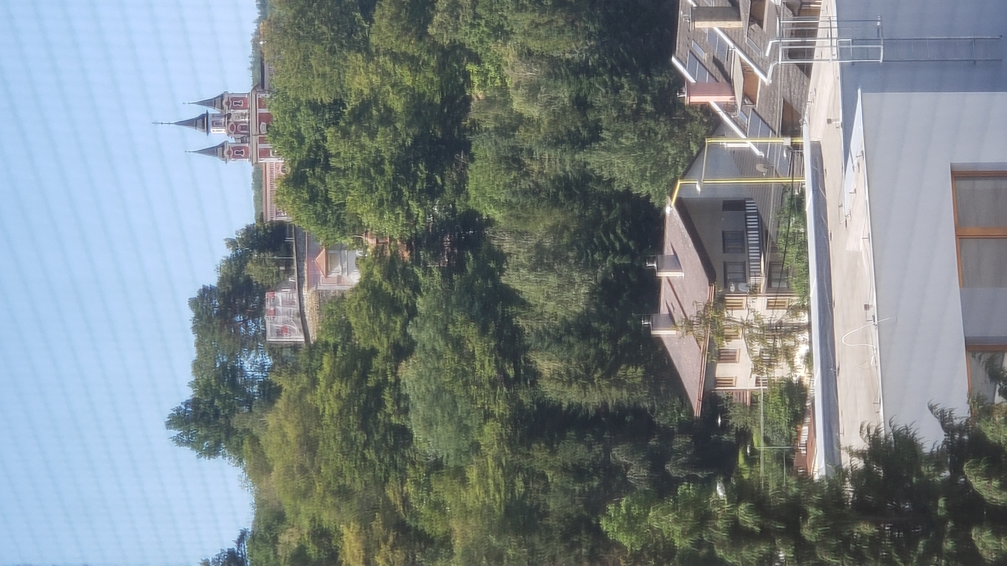 The image shows a hilly landscape with dense green trees and several buildings. In the background, on top of a hill, a red and white building with multiple spires, resembling a church or a castle, is visible. Below this, closer to the foreground, there are several residential buildings with brown roofs and visible windows. The foreground features the roof of a modern building with a ladder attached to its side. The sky is clear and blue. The scene appears to be a residential area on the outskirts of a city, possibly Prešov, Slovakia, given the architectural style and the natural environment. There are no people or animals visible in the image. The image is taken from a high vantage point, looking across the landscape.