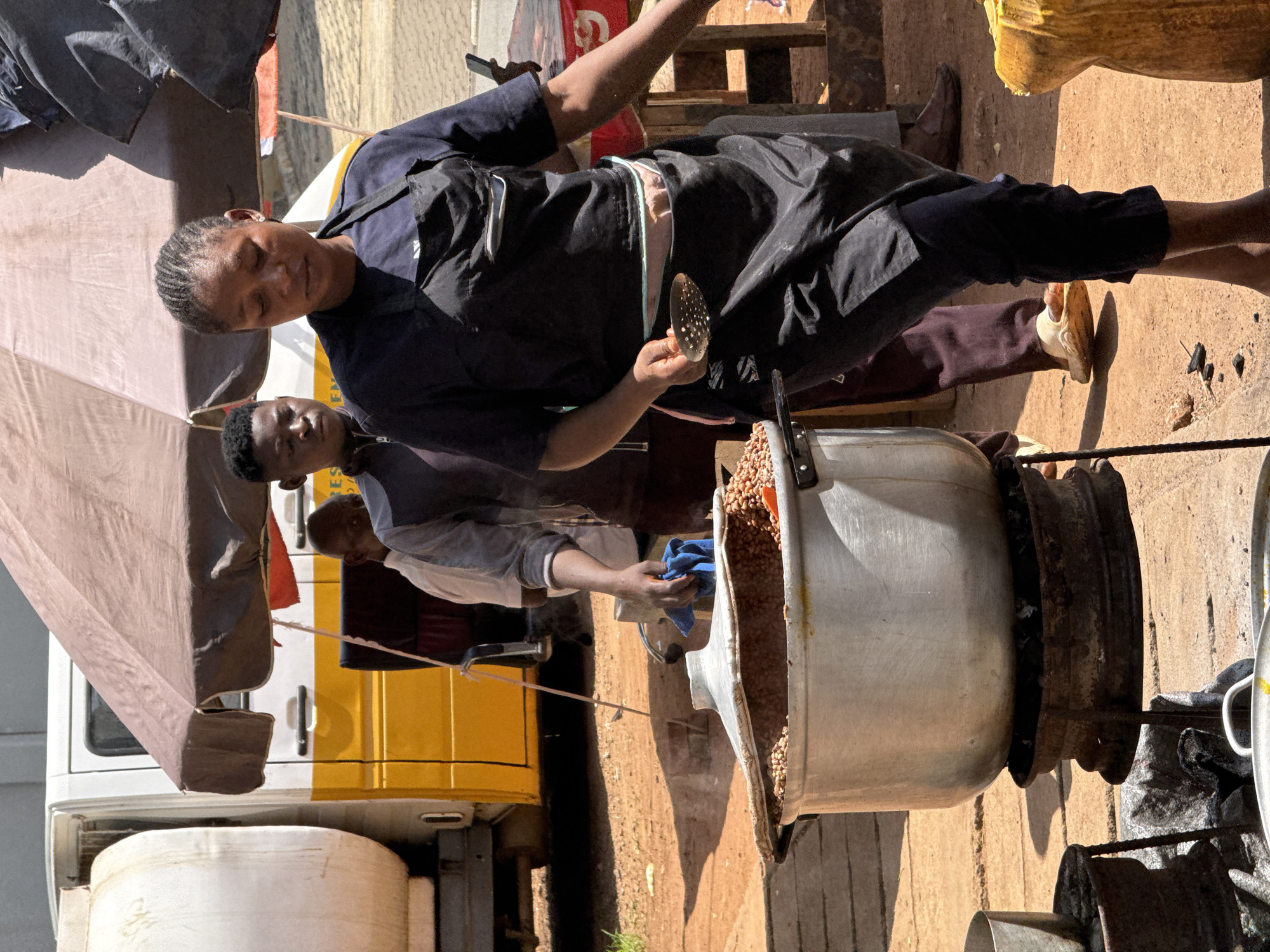 In Abuja, Nigeria, two individuals are preparing food outdoors. A woman, wearing a dark apron over casual attire, holds a perforated utensil, likely for straining or scooping. A younger man, also dressed casually, uses a blue cloth to handle a large metal pot filled with what appears to be peanuts or similar legumes. The pot sits atop a makeshift stove composed of metal rings, possibly repurposed tires or machinery parts. A white and yellow van is parked nearby, with a large umbrella extending from it to provide shade. A white container sits next to the van. The ground is dusty and sunlit.