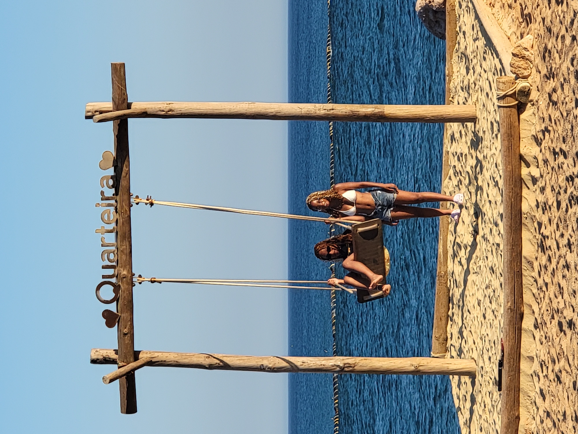 Two young women are positioned on a sandy area by the ocean, under a wooden swing set. The woman on the left is seated on the swing, which is made of dark wood and suspended by ropes. She is wearing sunglasses, a dark top, and has her legs and feet visible. The woman on the right is standing next to the swing set. She is wearing a white bikini top and denim shorts. Both women appear to be enjoying themselves. The swing set has a wooden beam across the top with the word "Quarteira" and two heart shapes displayed on it. The background consists of the blue ocean and a clear blue sky. The ground is covered in sand, with some wooden logs placed as a border.