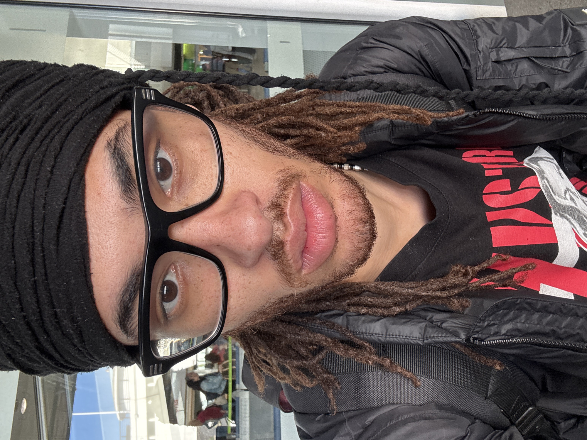 A close-up, rotated portrait shows a person with brown dreadlocks wearing black glasses and a black head covering. The person is also wearing a black jacket over a black t-shirt with red and white graphic elements. The background is a slightly out-of-focus urban setting, with hints of reflective glass and architectural features. The person appears to be indoors or near a window, as there is a reflection of people and the exterior of a building in the background. There is no indication that the image was taken in the city of San Bartolomé, Spain.