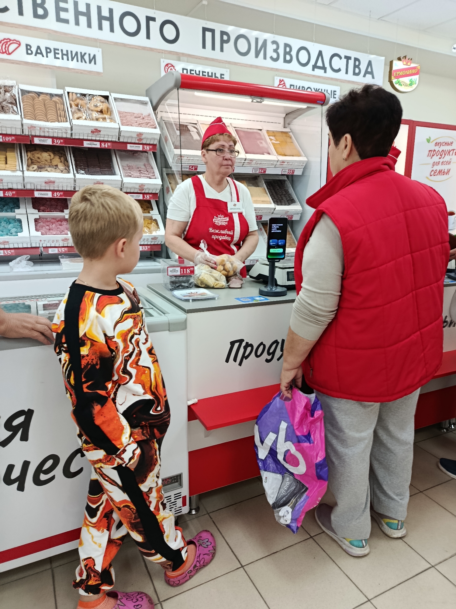 In a supermarket in Vereya, Russia, a young boy in a colorful tracksuit stands at a checkout counter. A female cashier in a red apron and cap is serving a woman wearing a red vest over a beige sweater. The woman is holding a pink and purple shopping bag. Behind the cashier are displays of various food items, including what appear to be cookies, candies, and packaged meats. A digital payment terminal is visible on the counter. The environment suggests a retail setting with product shelving and a tiled floor.