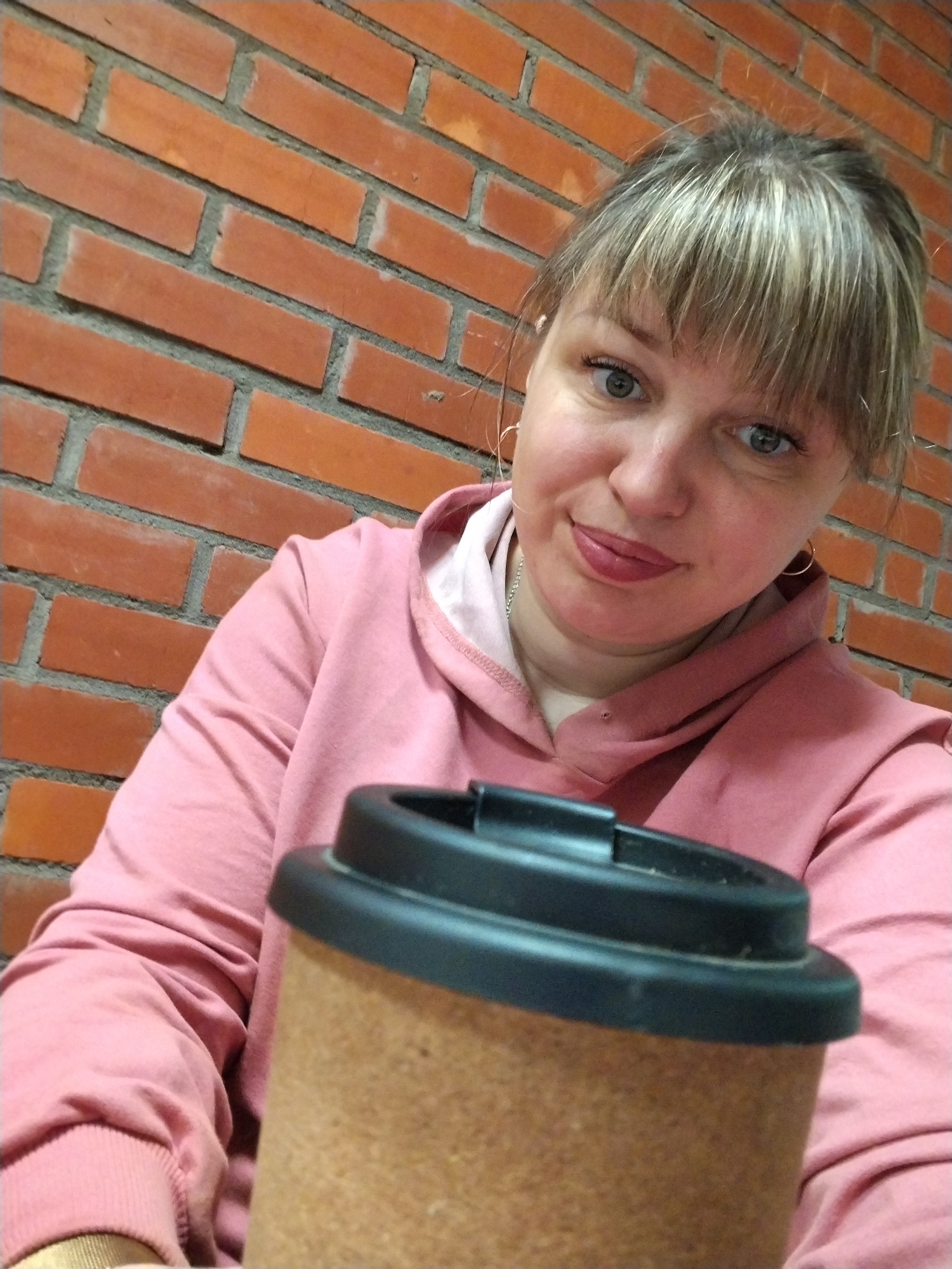 A close-up, eye-level portrait features a woman with fair skin, light-colored eyes, and light brown hair with bangs, which is styled upwards. She is wearing a pink hooded sweatshirt, small hoop earrings, and a thin necklace. Her lips are colored with red or pink lipstick, and she offers a faint, friendly smile directly at the viewer.
A travel mug is prominently placed in the foreground, partially obscuring her torso. The mug features a dark lid and a light brown, possibly cork-textured, body.
The background behind the woman is a brick wall, composed of reddish-orange bricks with grey mortar. The bricks are arranged in a stretcher bond pattern. The lighting appears soft and even, indicating an indoor setting. The woman's expression conveys a sense of contentment or amiability. No visible text is present anywhere in the image. This scene takes place in Rusanovo, Russia.