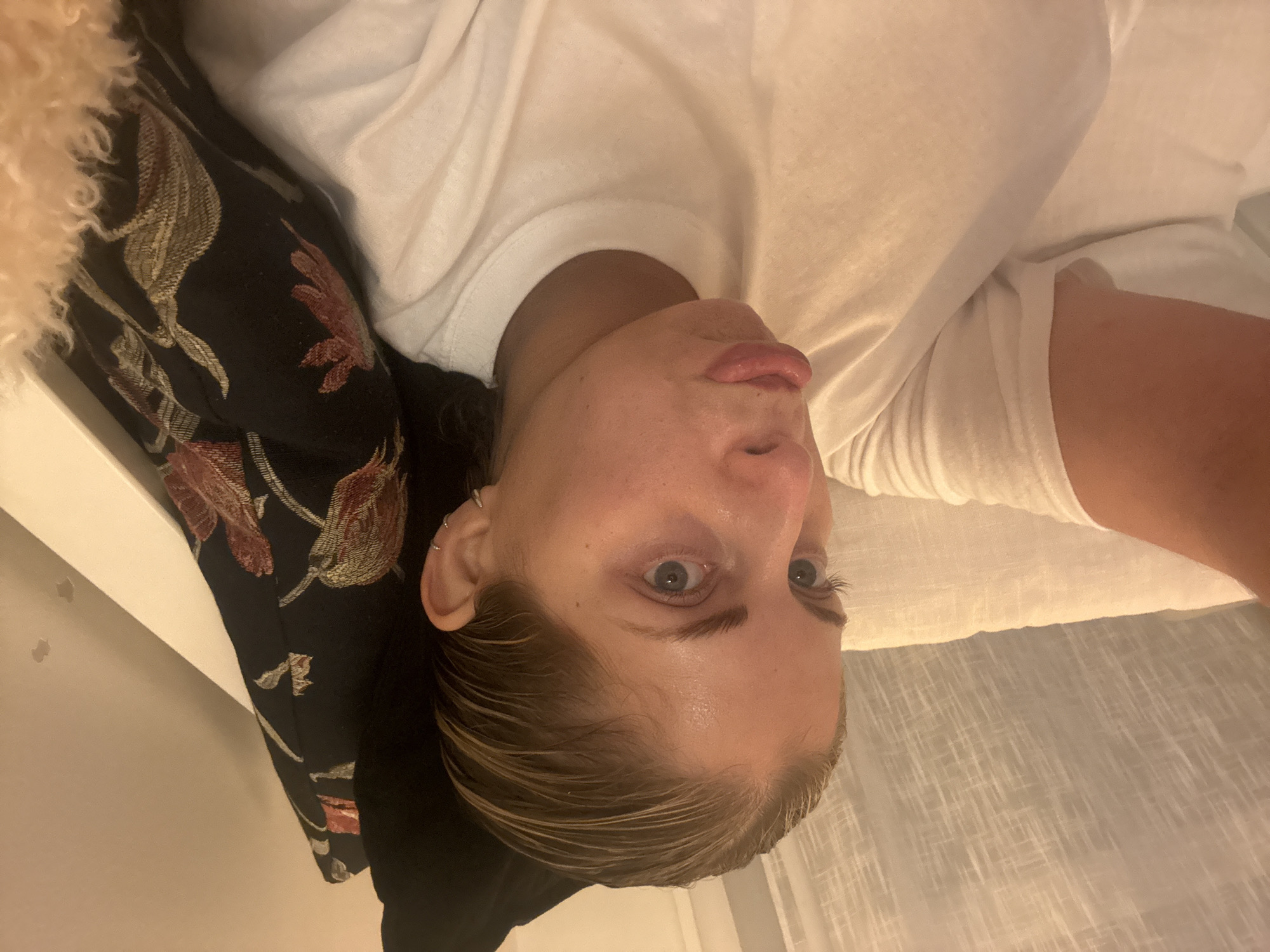 A fair-skinned person with blue eyes lies on a bed. The person wears a white shirt. A floral pillow sits next to their head. A white curtain hangs in the background. The location is not evident in the image, and it cannot be determined if the scene is in Paris, France.