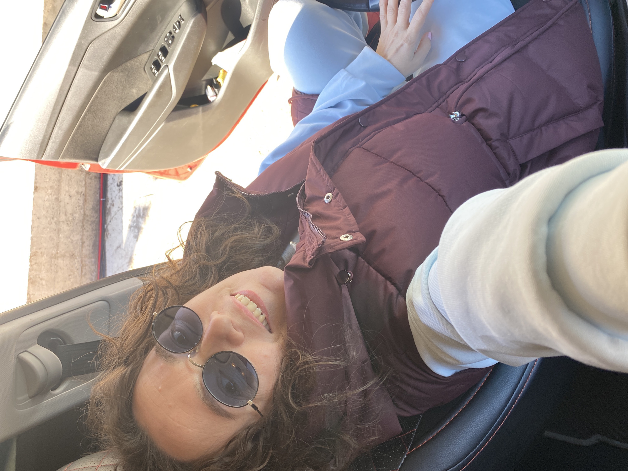 A person is sitting in the driver's seat of a red car. The person is wearing a maroon vest, light blue shirt, and round sunglasses. The car's interior is visible, including the door panel and steering wheel. The location is not identifiable as Ольховка, Russia, from the visual information provided.