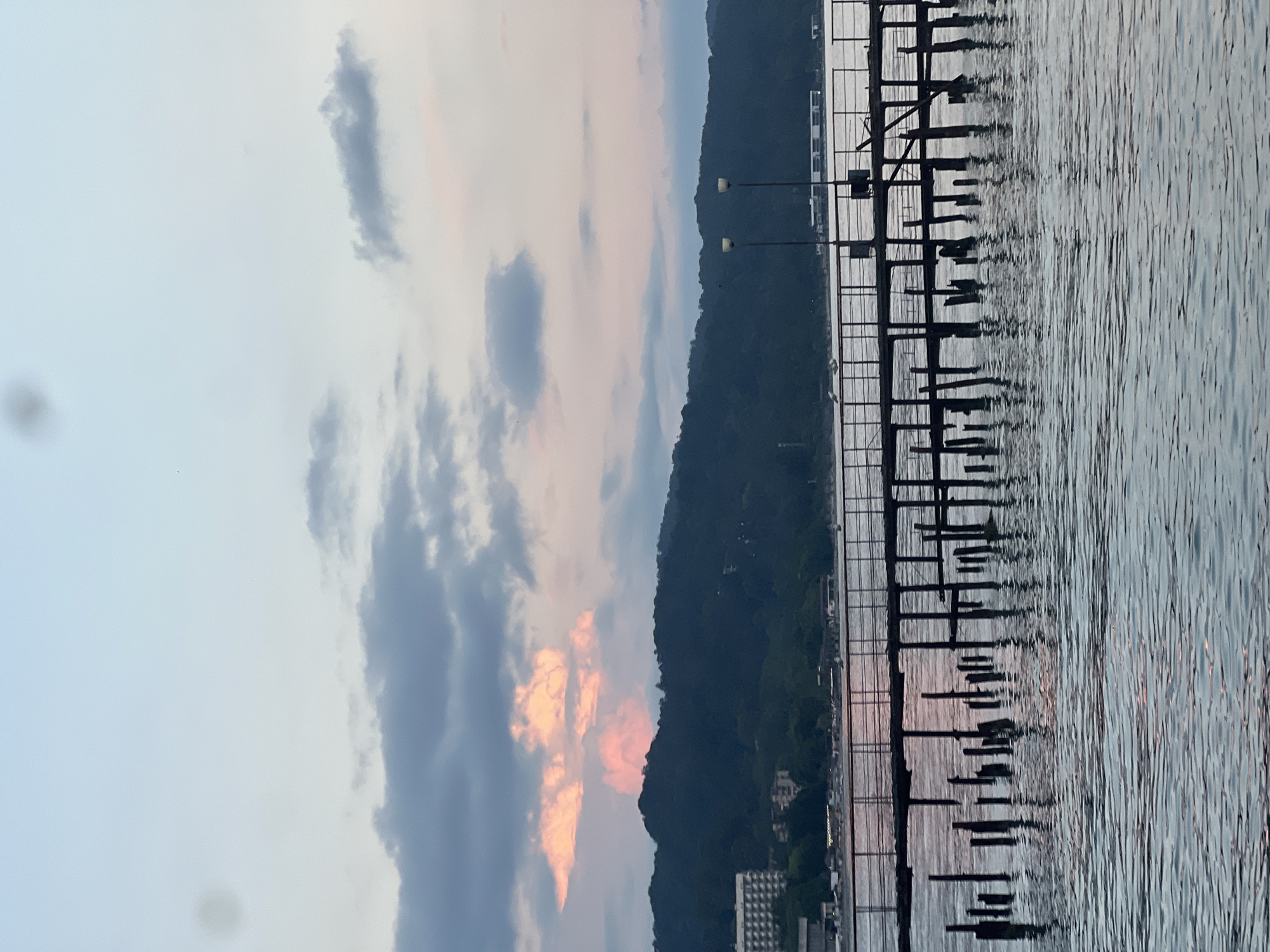 The image shows a serene waterfront scene at dusk. A dark, tree-covered mountain range dominates the background, with hints of buildings nestled at its base. In the foreground, a wooden pier or dock extends into the calm water, its pilings casting reflections that ripple with the gentle movement of the waves. The water itself is a muted grey-blue, with subtle highlights and shadows suggesting the changing light.
The sky is a canvas of soft hues, transitioning from a pale blue at the top to warmer, peachy tones near the horizon, where the sun has likely just set. Clouds are scattered across the sky; some are a soft grey, while others are tinged with the vibrant colors of sunset, creating a dramatic contrast against the dusky sky. Two streetlights with white lamps are visible on poles along the edge of the water, their illumination a subtle presence in the fading light.
The overall mood is peaceful and tranquil, evoking a sense of quiet contemplation. The scene appears to be in a natural setting, possibly a lake or a sheltered bay, given the calm water and the presence of the mountainous terrain. The time of day is clearly evening, as indicated by the sunset colors in the sky and the dim lighting. There is no visible text in the image.