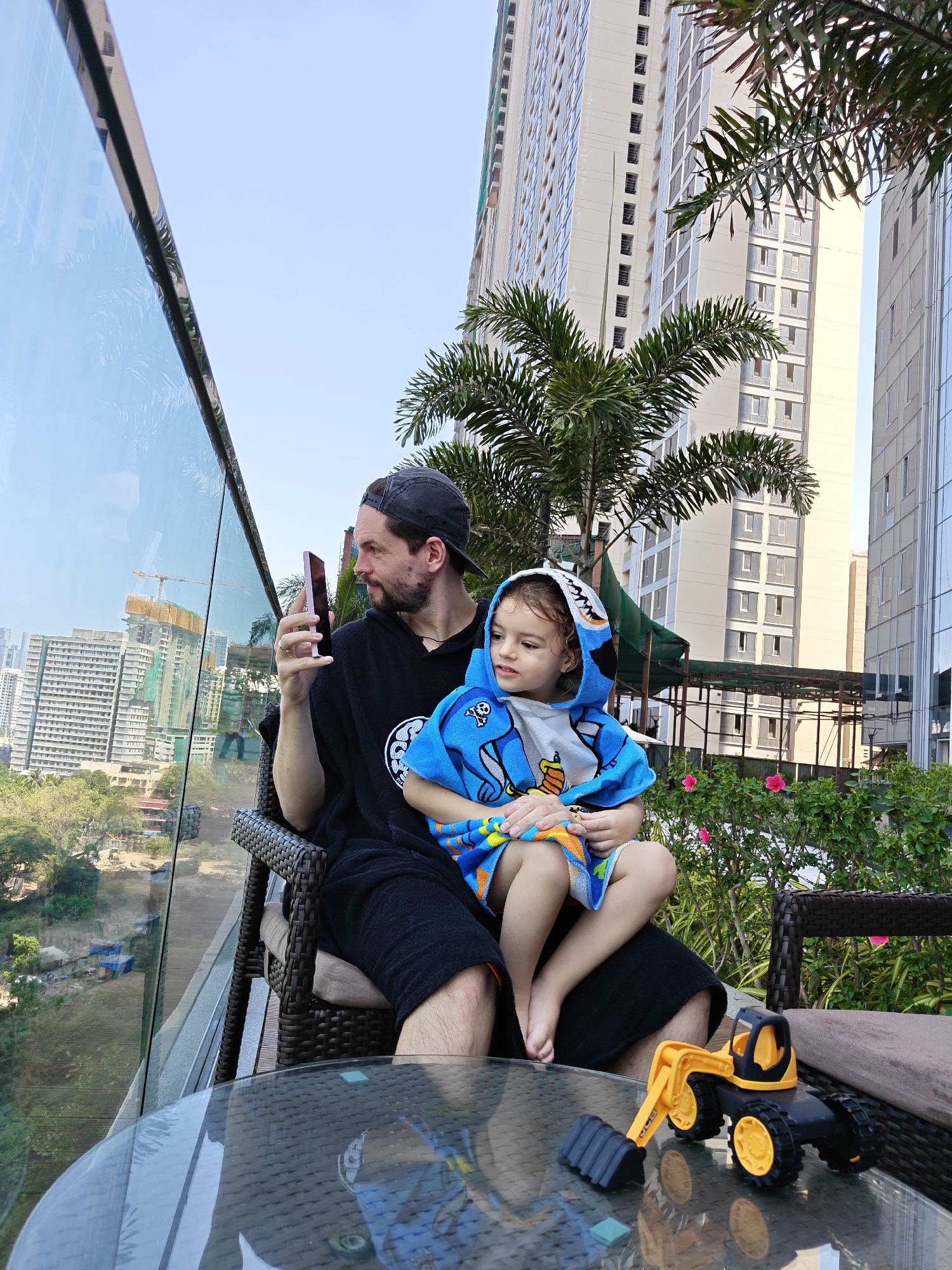 A male adult sits holding a young child on an outdoor high-rise terrace in Mumbai, India, overlooking an urban panorama. The man, with a beard and a dark baseball cap, wears a black hooded garment and is seated in a dark wicker chair. He holds a pink smartphone in his left hand, looking at its screen. A light-skinned child with curly hair sits on his lap, wrapped in a blue hooded towel featuring a shark design, looking towards the left foreground.
In the foreground, a round glass-top table with a dark wicker base displays a yellow and black toy excavator. Behind the man and child, a clear glass railing with a dark frame separates the terrace from the city view. The terrace features tropical vegetation, including palm trees and bushes with pink flowers.
The background reveals a dense urban skyline dominated by numerous high-rise buildings, many appearing residential or commercial, under a clear sky. Several buildings are visibly under construction, marked by scaffolding, green netting, and multiple tower cranes. One prominent crane is visible through the glass railing to the left. The terrain directly below the terrace appears less developed, showing patches of dirt and green foliage.