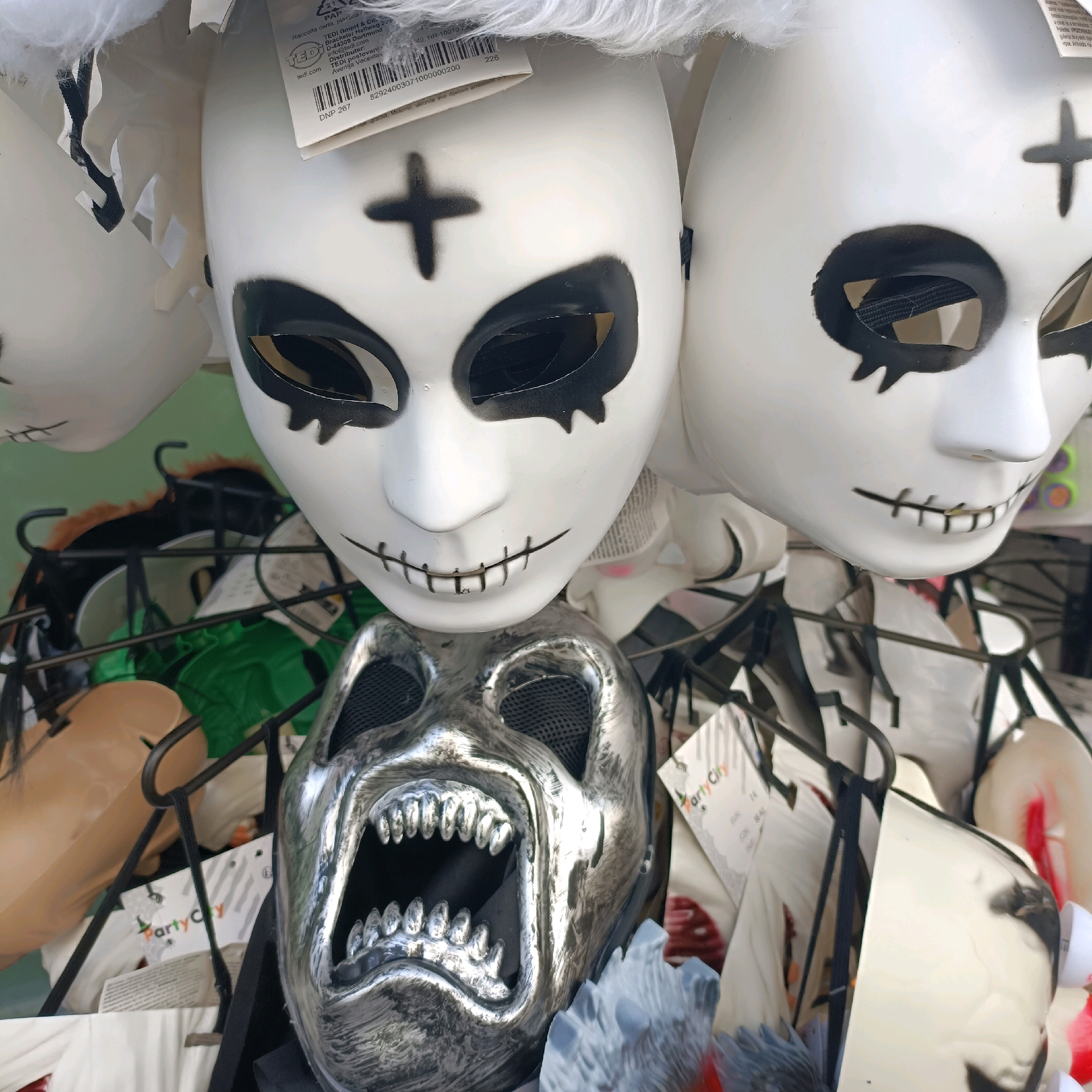 This is a close-up, eye-level shot of a display of Halloween masks. In the foreground and center, a silver, metallic mask with a gaping mouth full of sharp teeth is prominently featured. To the right and behind it, several white masks with black markings are visible. One mask has a black cross above the eyes and stitched mouth detail, another shows similar stitched mouth detail. In the background, more masks and assorted merchandise are visible, some hanging on metal racks. Partially obscured tags, one labeled "Party City," suggest this is likely a retail display. The lighting is bright and diffused, likely from an indoor store environment. There are no people or active interactions depicted. The scene is static and focused on the product display. The location is identified as Offenbach am Main, Germany.