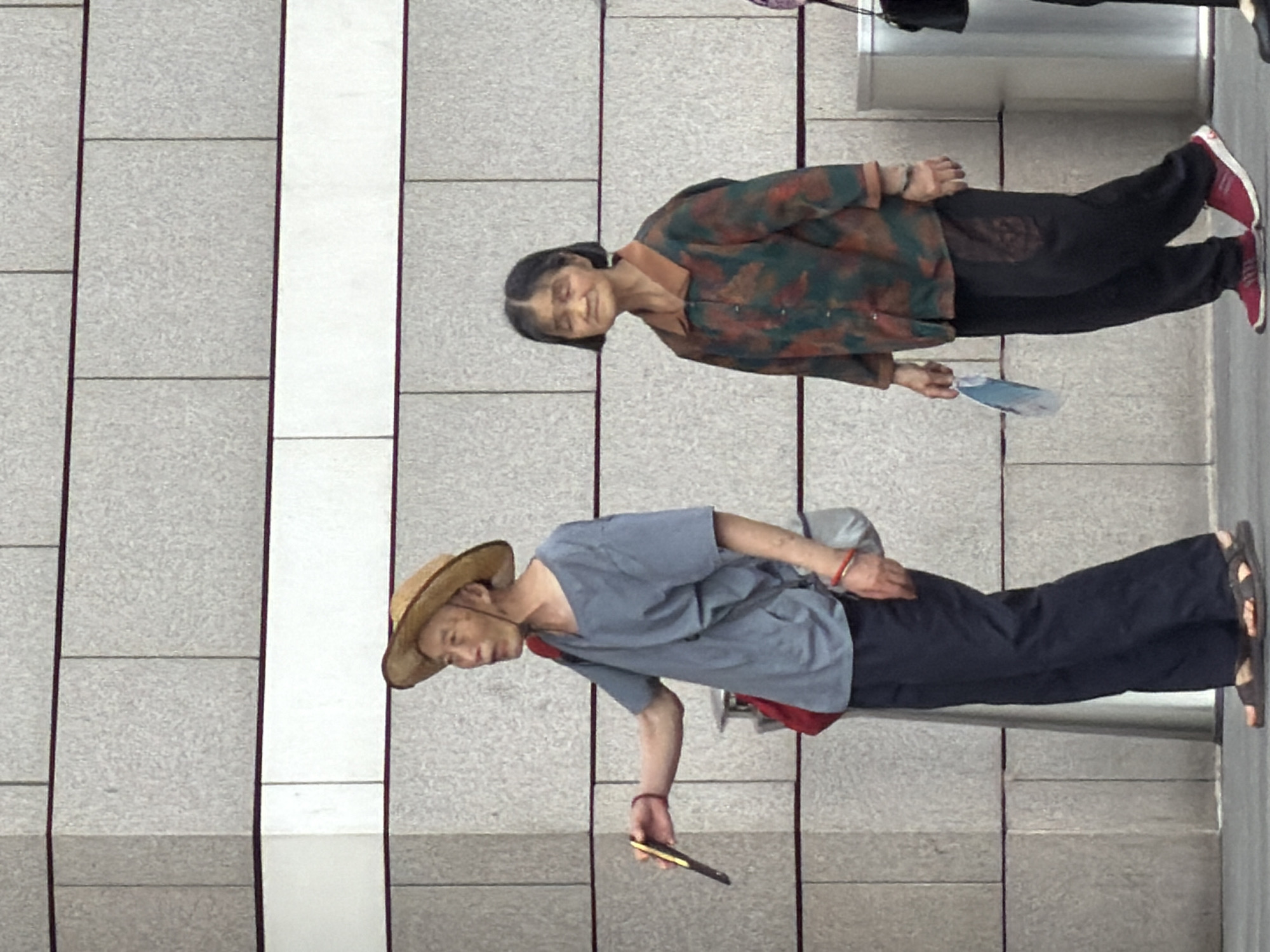 Two elderly individuals, a man and a woman, are walking in front of a textured, grey brick building in Yuzhong District, China. The man, on the left, wears a straw hat, a loose blue t-shirt, and dark pants, carrying a red bag over his shoulder and a small fan in his right hand. The woman, on the right, has short black hair, wears a patterned button-up shirt and dark pants, and holds a blue and white fan in her left hand. Both are wearing sandals. The environment appears to be an outdoor urban setting with a paved walkway.