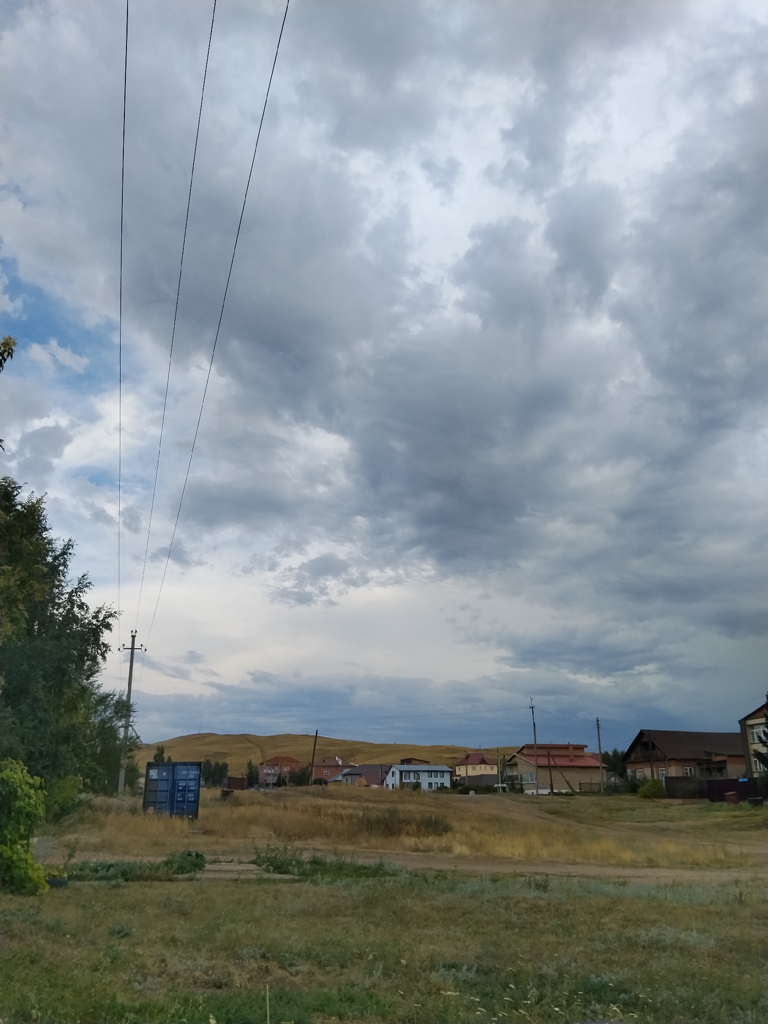 The image captures a rural or semi-rural scene under a dramatic, cloudy sky. In the foreground, dry grass and some patches of green vegetation cover the ground. A dirt path or road runs horizontally across the mid-ground. To the left, a large blue shipping container stands prominently. Behind the container, a utility pole with wires is visible, along with the lush green foliage of trees. In the background, a cluster of houses with various roof colors, including red and brown, are situated on a gentle, rolling hill. The sky is filled with heavy, gray clouds, suggesting an overcast or potentially stormy day. The landscape appears to be located in the outskirts of Kokshetau, Kazakhstan, characterized by its open spaces and dispersed residential areas. There are no people or animals visible in the frame.