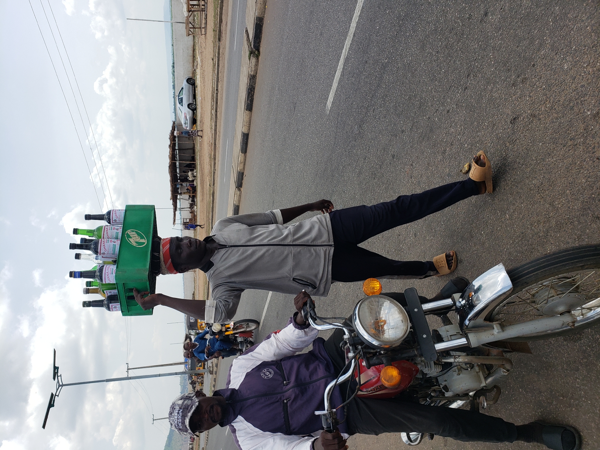 This image, rotated 90 degrees clockwise, captures a dynamic daytime street scene in Lokoja, Nigeria. The main subjects are two men and a motorcycle on an asphalt road.
In the immediate foreground, a man wearing a light grey zip-up jacket and dark trousers walks alongside a red and white motorcycle. He expertly balances a green plastic crate on his head, which is filled with multiple glass bottles. The bottles include both clear and dark varieties, with labels clearly visible, predominantly displaying "GHANA HERBAL." The green crate itself features a "7up" logo. This man is wearing light brown sandals.
Seated on the red and white motorcycle, holding the handlebars, is another man. He is dressed in a purple and white jacket and a patterned cap. Text on his cap appears to read "MODERN DESIGN." The motorcycle has chrome handlebars, a large round headlight, and orange turn signals.
The background reveals more of the street environment. The asphalt road has a single white line marking and a black and white painted curb. Beyond the curb, there's a dirt shoulder with simple wooden structures, likely stalls, and several goats are visible in this area. A white car is parked further down the road. Utility poles with overhead wires stretch across the scene against a bright, cloudy sky, indicating clear weather. The overall activity suggests a typical day of commerce and movement on a local thoroughfare.