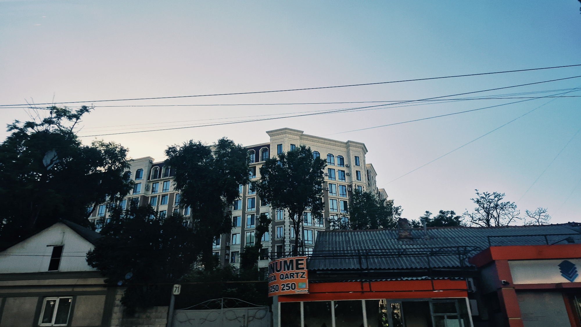 The image captures a street view in Chișinău, Moldova, likely during twilight, as indicated by the soft, gradient sky transitioning from light yellow near the horizon to a pale blue. Dominating the scene is a tall, modern apartment building with a light beige facade, numerous blue-tinted windows, and decorative architectural elements at its crown. In the foreground, a smaller, older building with a grey, corrugated metal roof is visible, featuring an orange awning with signage that reads "NUME GRICEA QARTZ" followed by prices "250 250". To the left of this, another low-rise structure with white walls and a dark roofline is partially visible, with a window on its upper level. Dense foliage, consisting of dark green trees, partially obscures the lower portions of the apartment building and the older structures, suggesting a mix of urban development and greenery. Several overhead power lines crisscross the sky, casting faint shadows. A sign with the number "71" is present on a pole to the right of the white building. The overall impression is a typical urban residential area with a blend of older and newer construction. No people are visible in the frame.