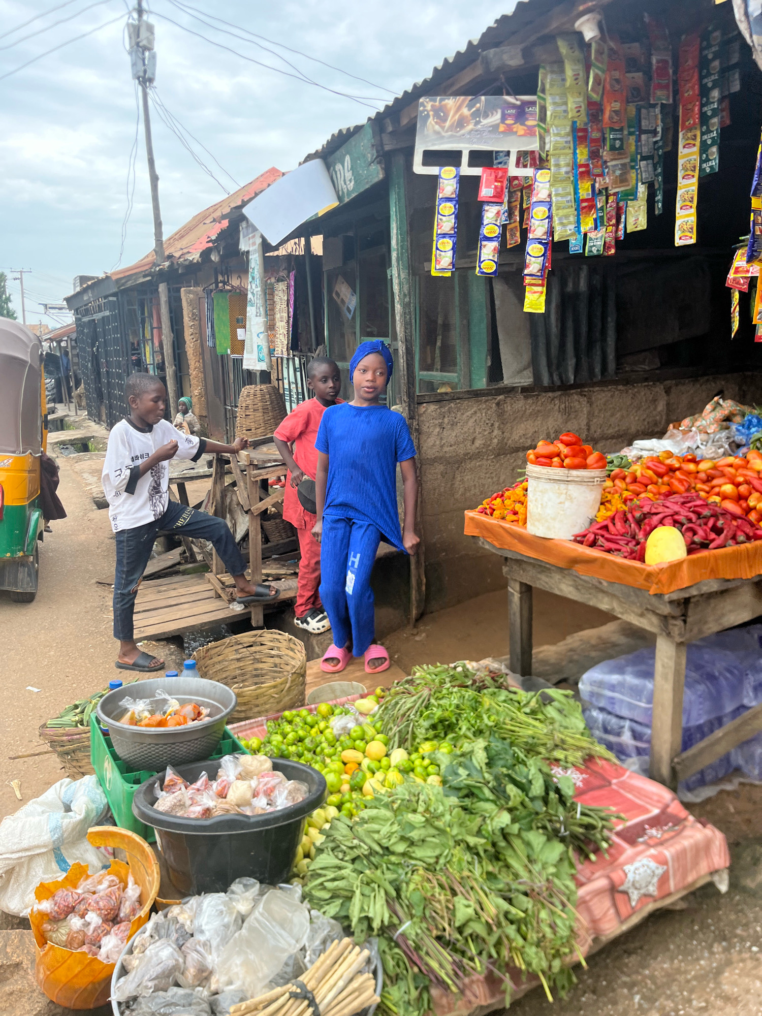 In Jos, Nigeria, a young Black boy in a white t-shirt and dark pants stands on a wooden platform at a roadside market, holding a small white object to his mouth. To his right, a younger Black boy in a red outfit stands on a similar platform, looking towards the camera. Another Black girl in a bright blue outfit with a headscarf stands on the ground to the right of the boy in red, also looking towards the camera. Behind them, a stall displays various packaged goods, and a table in front of the girl is laden with a colorful assortment of fresh produce, including tomatoes, chili peppers, and leafy greens. An auto-rickshaw is partially visible on the left. The scene captures the vibrant atmosphere of a bustling market in Jos.