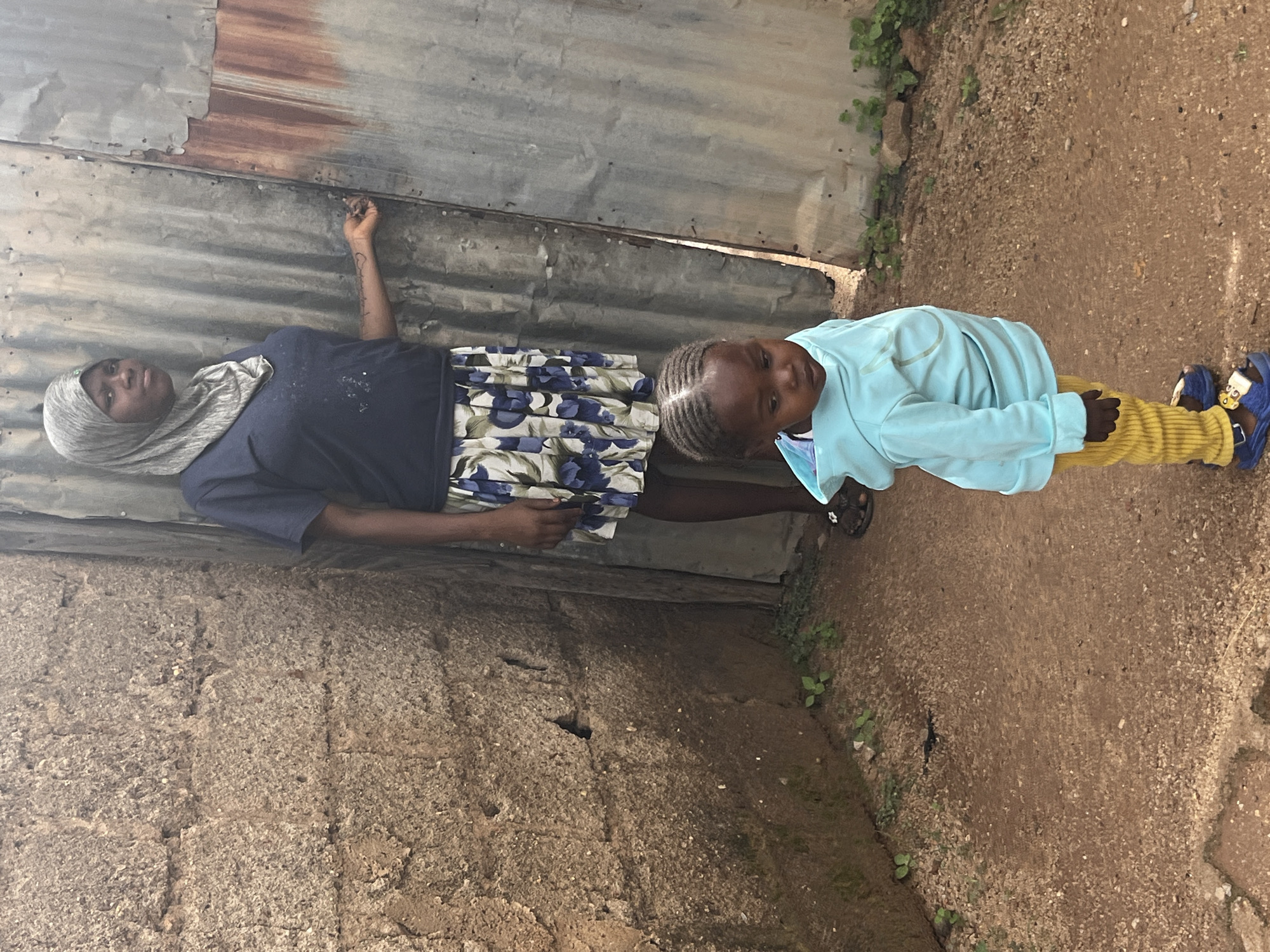 In an outdoor setting, two individuals, an adult and a child, are captured in a vertical, candid photograph. The adult, on the left, wears a dark blue t-shirt and a light grey hijab, with a floral skirt featuring blue and white patterns. The child, to the right, is dressed in a light blue long-sleeved top and yellow ribbed trousers.
They appear to be standing next to a weathered, corrugated metal wall, which shows signs of rust and age. In front of them, a rough, gravelly ground leads away from the wall. The overall scene suggests a humble, possibly rural or peri-urban environment, consistent with the provided location context of Jos, Nigeria.
The individuals are not actively engaged in a specific event but seem to be posing or standing casually for the photo. The adult's gaze is directed slightly upwards and to the left, with a neutral expression, while the child looks directly at the camera with a more engaged expression.
Notable details include the textures of the metal wall and the rough ground, as well as the contrasting colors of their clothing. The lighting suggests it could be daytime, with no strong indications of specific weather conditions like rain or intense sun, beyond general daylight. There is no visible text in the image. The composition, with the subjects framed by the wall and ground, gives a sense of their immediate surroundings.
