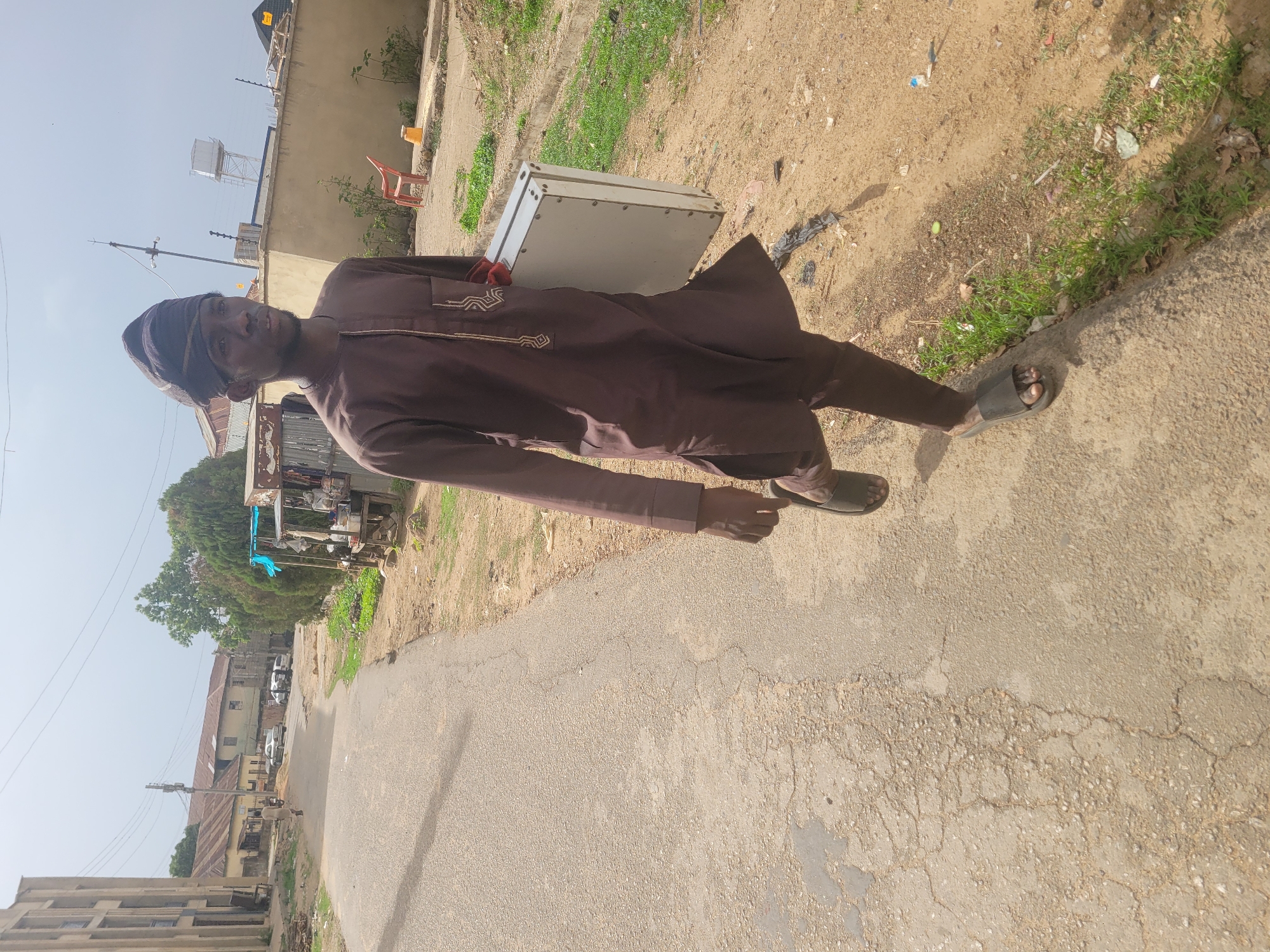 A full-body shot of a slender man in traditional Nigerian attire, walking along a cracked asphalt road in Jos, Nigeria. He is wearing a brown, long-sleeved agbada with light-colored embroidery around the neck and chest, matching brown trousers, and open-toed sandals. A head wrap, dark in color, is also worn. He carries a gray metal briefcase or box. The surrounding environment includes residential buildings, some with corrugated iron roofs, and trees, suggesting an urban area of Jos. The lighting indicates a sunny day.