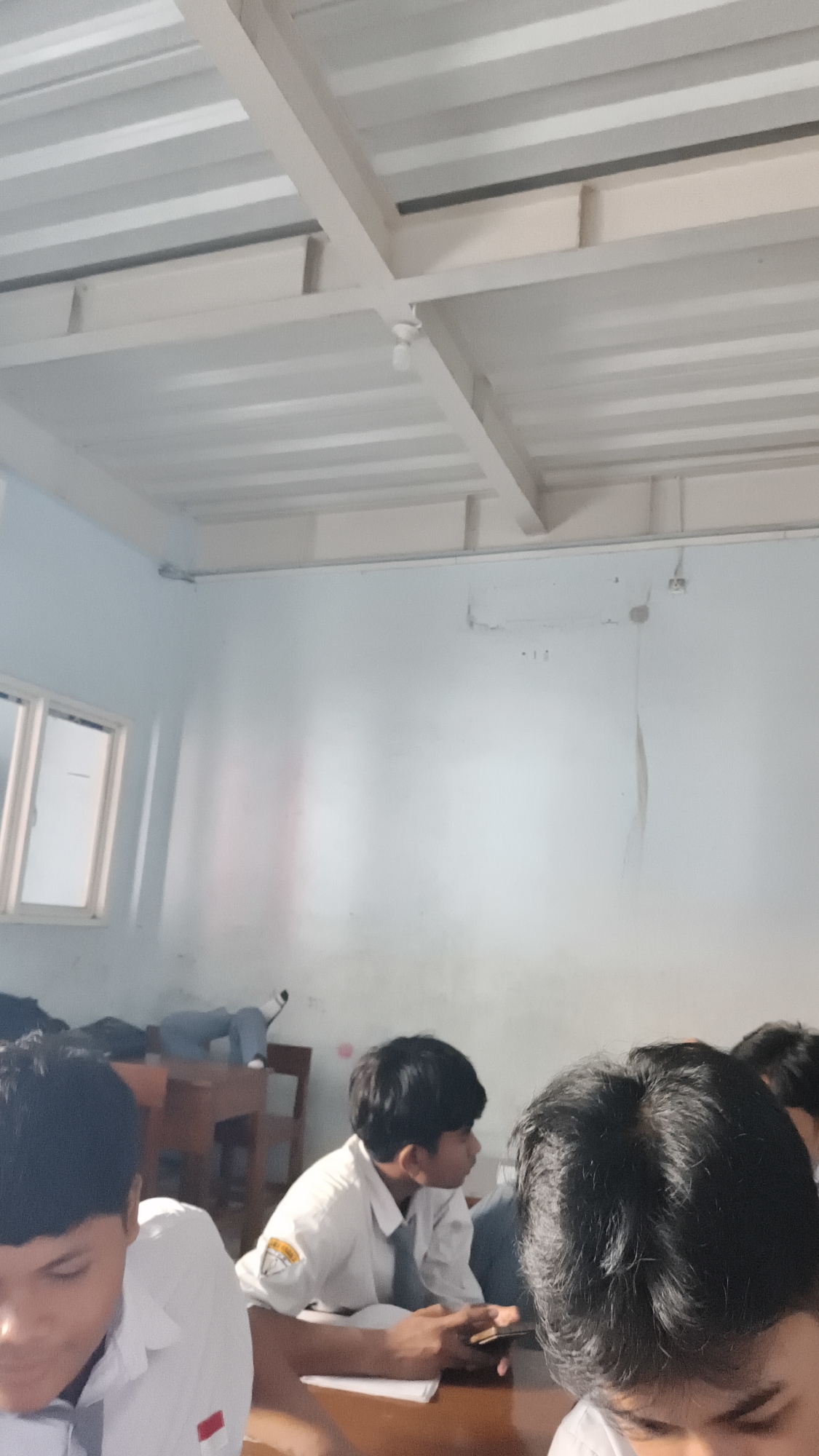 The image shows an indoor scene, likely a classroom, with a white ceiling and light blue walls. In the foreground, the heads and shoulders of several young males wearing white school uniforms are visible. One boy in the center, wearing a white shirt and grey tie, is looking to his right and holding a mobile phone. He is seated at a wooden desk. Another boy in the left foreground is also wearing a white school shirt. In the background, a window with white frames is visible on the left wall, and a desk with a person lying on it can be seen. The overall impression is that of a school setting in Katrungan, Indonesia, possibly during a class break or a less formal learning session.