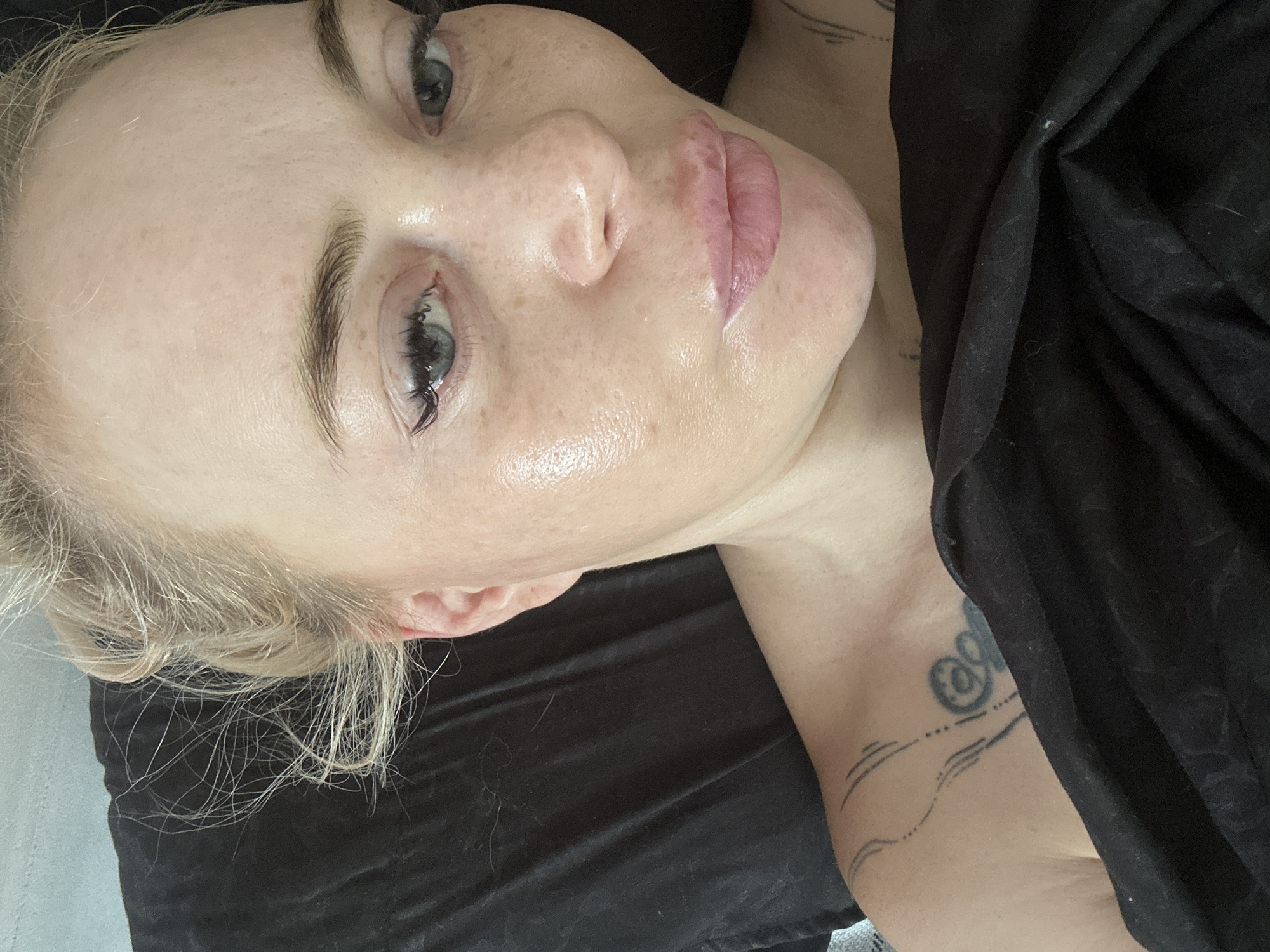 A close-up, overhead shot shows a blonde woman with light blue eyes lying down, her head tilted slightly upwards. She is wearing makeup, including eyeliner, and has freckles across her nose and cheeks. Her hair is pulled back loosely, with wisps framing her face. Her lips are slightly parted and appear to be glossed. The background is a dark, crumpled fabric, possibly a blanket or sheet. A tattoo is visible on her left shoulder and upper chest area. The lighting is bright, creating a slight sheen on her skin.