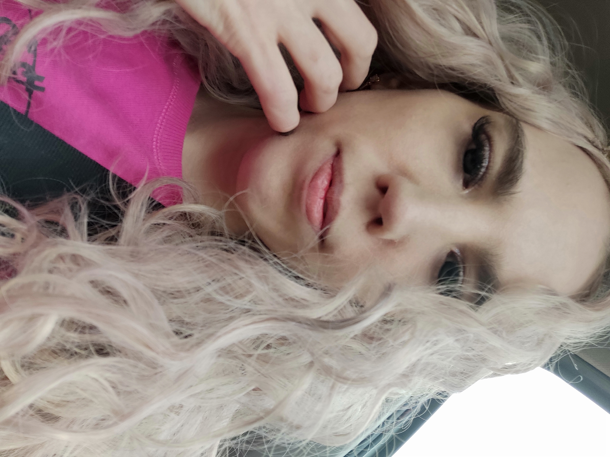 A close-up, selfie-style photograph shows a woman indoors, her face at an oblique angle towards the camera. She has long, wavy, blonde hair that partially obscures her face. Her skin appears fair, and she wears eye makeup including visible eyelashes. Her lips are lightly colored. Her left hand touches her face near her ear. She wears a bright pink and black sweatshirt. The overall lighting suggests an indoor setting. The location is unknown.