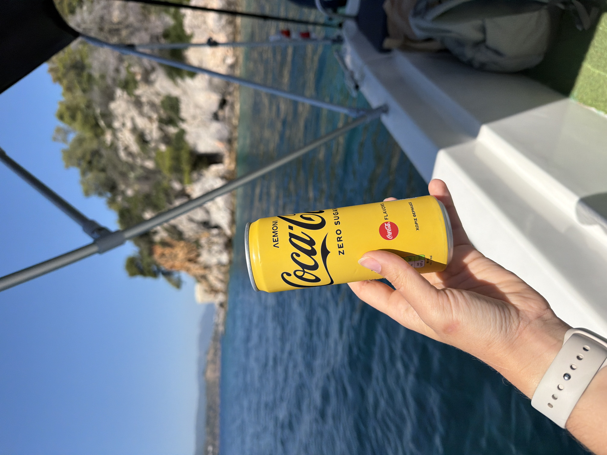 The image, taken with a vertical orientation, captures a right hand holding a vibrant yellow Coca-Cola Zero Sugar can against a bright, sunny marine backdrop. The scene is set on a boat, with the blue sea and a rugged coastline visible in the background, consistent with a location like Poros, Greece.
The Coca-Cola can is the central object, prominently displaying its red and black "Coca-Cola" logo, with "ZERO SUGAR" and "ΛΕΜΟΝΙ" (Lemon) printed in black on its yellow surface. Other visible Greek text on the can includes "ΧΩΡΙΣ ΘΕΡΜΙΔΕΣ" (Calorie-free) and "FLAVORS," along with partial nutritional information.
The hand holding the can has manicured nails and wears a white Apple Watch with a patterned strap on the wrist. A faint reddish mark is visible on the back of the hand.
The setting is clearly a boat, with parts of a white hull and metal railings visible in the foreground. Beyond the boat, the water is a clear, deep blue, with subtle ripples. In the midground and background, a rocky coastline or island is visible, covered sparsely with green Mediterranean vegetation under a cloudless, bright blue sky.
The strong sunlight indicates daytime and good weather, suitable for boating and enjoying a cold drink. The activity is simply holding a beverage while on a boat in a scenic location.