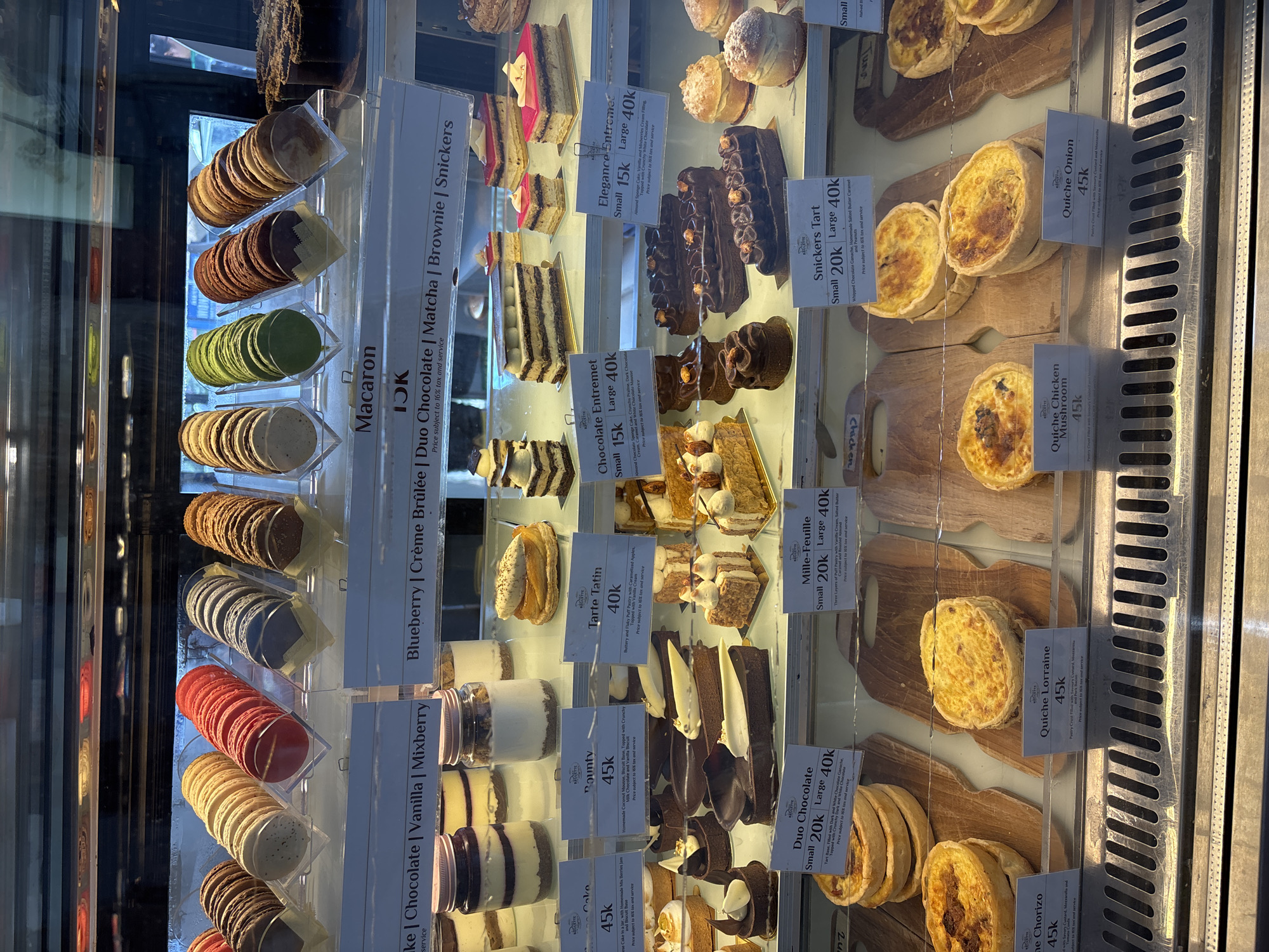 The image shows a display case filled with various pastries and desserts, likely in a shop located in Denpasar, Indonesia, as indicated by the currency "k" (thousands of Indonesian Rupiah). The case contains macarons in multiple colors, cakes, tarts, and quiches. Price tags are visible for each item. The macarons are arranged in rows with multiple flavors. The cakes include chocolate, vanilla, mixed berry, blueberry, crème brûlée, matcha, brownie, and snickers flavors. The tarts are Duo Chocolate, Mille-Feuille, and Snickers Tart. Quiche varieties are Lorraine, chicken mushroom, and onion. The interior of the shop appears modern, with the pastries neatly organized in the display case. The shot is taken close up.