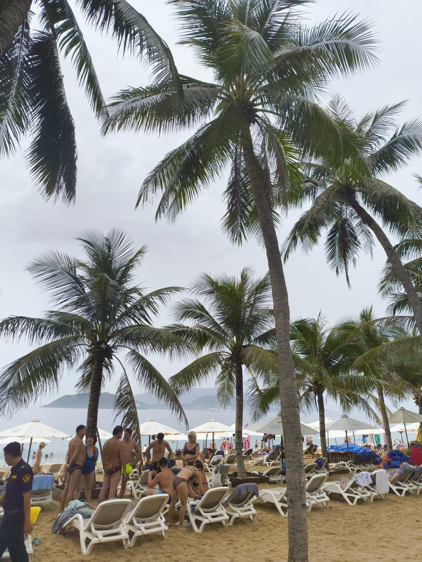 The image depicts a crowded beach scene in Nha Trang, Vietnam, under an overcast sky. Tall palm trees dominate the foreground and midground, their fronds casting shadows on the sand. The beach is lined with rows of white lounge chairs, many occupied by people sunbathing or relaxing.
In the midground, several groups of people are visible. A cluster of individuals stands near some lounge chairs, some in swimsuits, appearing to converse. To the left, a person in a dark uniform, possibly a security guard or staff member, walks towards the viewer. Further back, several white beach umbrellas are set up, providing shade.
In the background, the calm sea is visible with distant hills or mountains on the horizon. Several boats are scattered on the water. The overall atmosphere is one of a busy tourist beach. The presence of numerous lounge chairs, umbrellas, and people in swimwear suggests a resort or public beach environment.