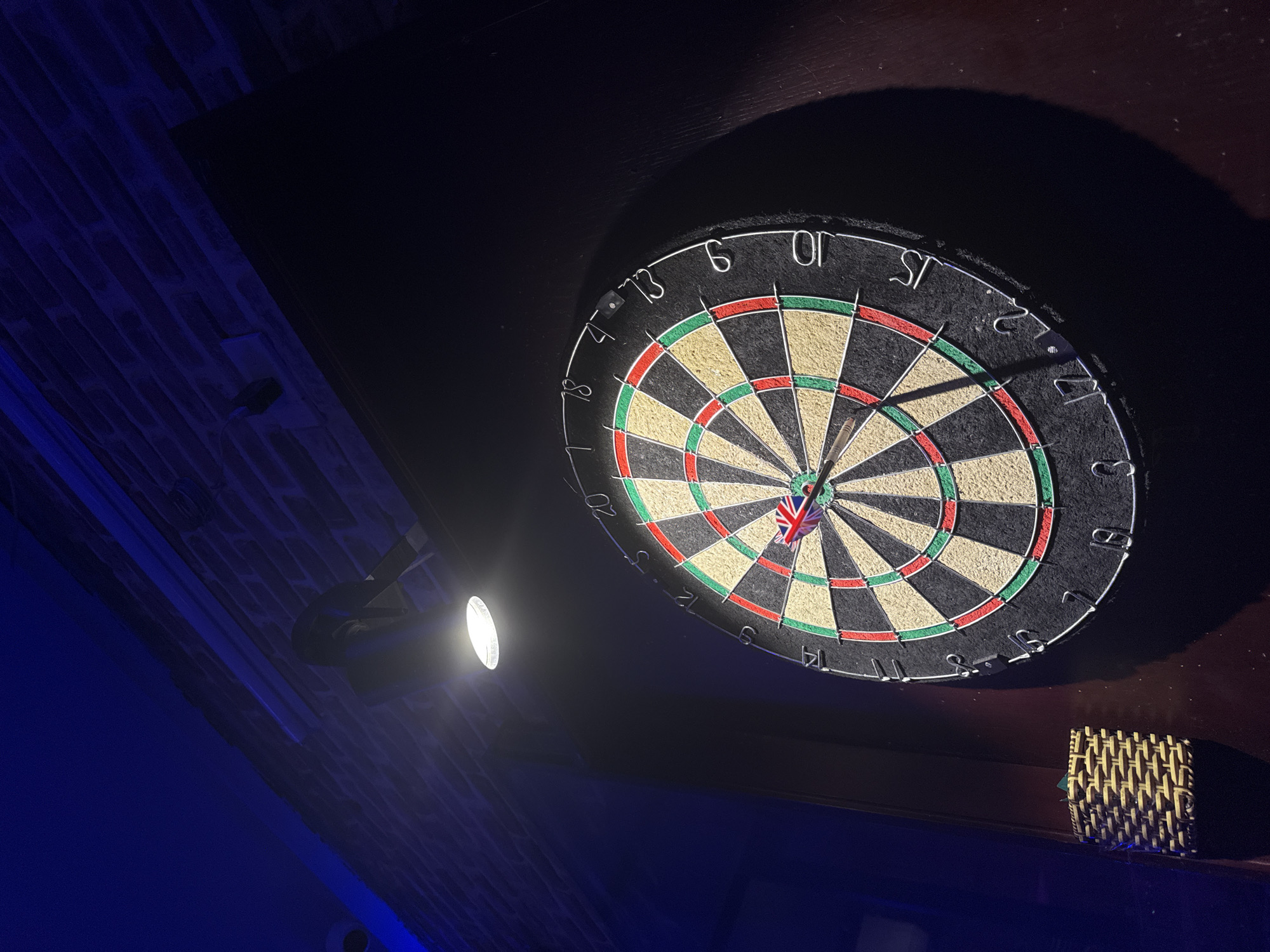 An indoor, low-light photograph captures a dartboard prominently displayed on a dark surface, likely a wall or cabinet. A single dart, adorned with a Union Jack flag on its flight, is embedded near the center of the board, suggesting an ongoing or recently played game. The dartboard itself is a standard bristle type, with numbers (such as 6, 10, 13, 15, 17, 18, 20) marking the scoring sections around its perimeter.
A bright, circular spotlight illuminates the dartboard from the lower-left, casting distinct shadows and highlighting the texture of the board. To the left of the dartboard, a brick wall is visible, bathed in a deep blue ambient light, contrasting with the warm glow on the dartboard. The lower-left portion of the frame is dominated by this intense blue hue, possibly from decorative lighting. In the lower-right, a small, rectangular object with a woven texture, resembling a coaster holder or a small basket, sits on the dark surface next to the dartboard.
The overall setting appears to be an entertainment space such as a bar, pub, or a home game room in Đà Nẵng, Vietnam, at night or in the evening, given the dim ambient light and targeted illumination. The blue lighting adds a cool, modern atmosphere to the scene.