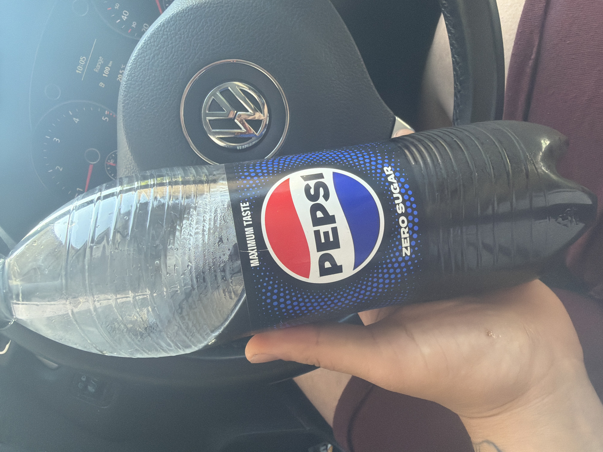 The image depicts a close-up view from inside a Volkswagen car, likely in Shumen, Bulgaria, featuring a person's hand holding a bottle of Pepsi Zero Sugar. The bottle, partially filled with a dark liquid and covered in condensation, indicates it is cold. Its label is prominently displayed, featuring the classic red, white, and blue Pepsi logo against a black background with blue dots, alongside text that reads "PEPSI", "ZERO SUGAR", and "MAXIMUM TASTE".
The person's hand, visible in the foreground, shows a small blemish or mole on the back of the wrist area. A glimpse of maroon-colored clothing can be seen in the background.
The setting is a car interior, with a dark grey Volkswagen steering wheel prominently featured, bearing its metallic logo. Behind the steering wheel, parts of the instrument cluster are visible, displaying key information. The digital screen shows the time as "10:05", an outside temperature of "20.5°C", and a remaining "Range" of "100 km". Analogue speedometer and tachometer gauges are also partially visible.
The lighting suggests a bright, sunny daytime, consistent with the visible temperature. The scene captures a casual moment of refreshment inside a vehicle.