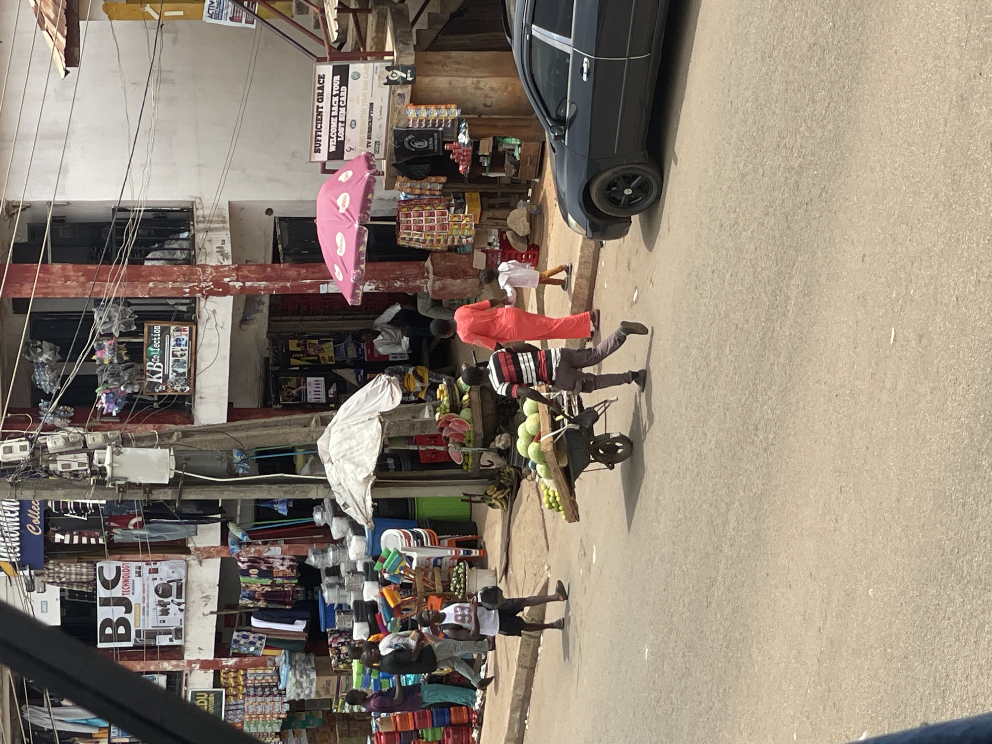 The image shows a street scene in Bukuru, Nigeria. A man in an orange outfit walks past a parked car. Another man pushes a wheelbarrow loaded with green coconuts. People stroll past shops selling clothing, kitchenware, and various other goods under umbrellas. Power lines crisscross the street above. The buildings appear to be multiple stories with various advertisements and signs, including one reading "Sufficient Grace."