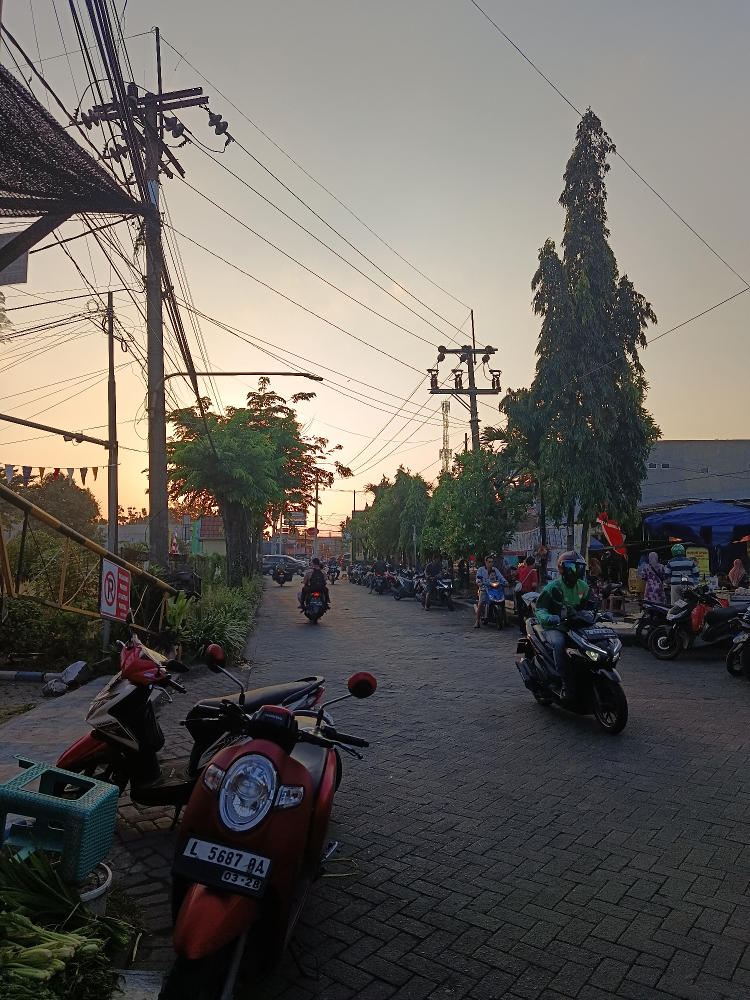 The image depicts a street scene in Keboansikep, Indonesia, likely during the late afternoon or early evening as indicated by the warm, low-angled sunlight. The road is paved with interlocking bricks. A multitude of motorcycles are parked along the sides of the road and in use, with several individuals riding them. One motorcycle in the foreground is red with a license plate reading "L 5687 BA 03.28". To the left, a green plastic basket sits on the ground next to a pile of green vegetables. Overhead, a complex network of electrical wires and poles dominates the sky. Trees line the side of the road, some appearing tall and slender, others more bushy. Buildings, some with awnings and stalls, are visible in the background. Several people are present, some on motorcycles and others appearing to be walking or standing near the stalls. The overall impression is a busy, active street in a semi-urban or market area.