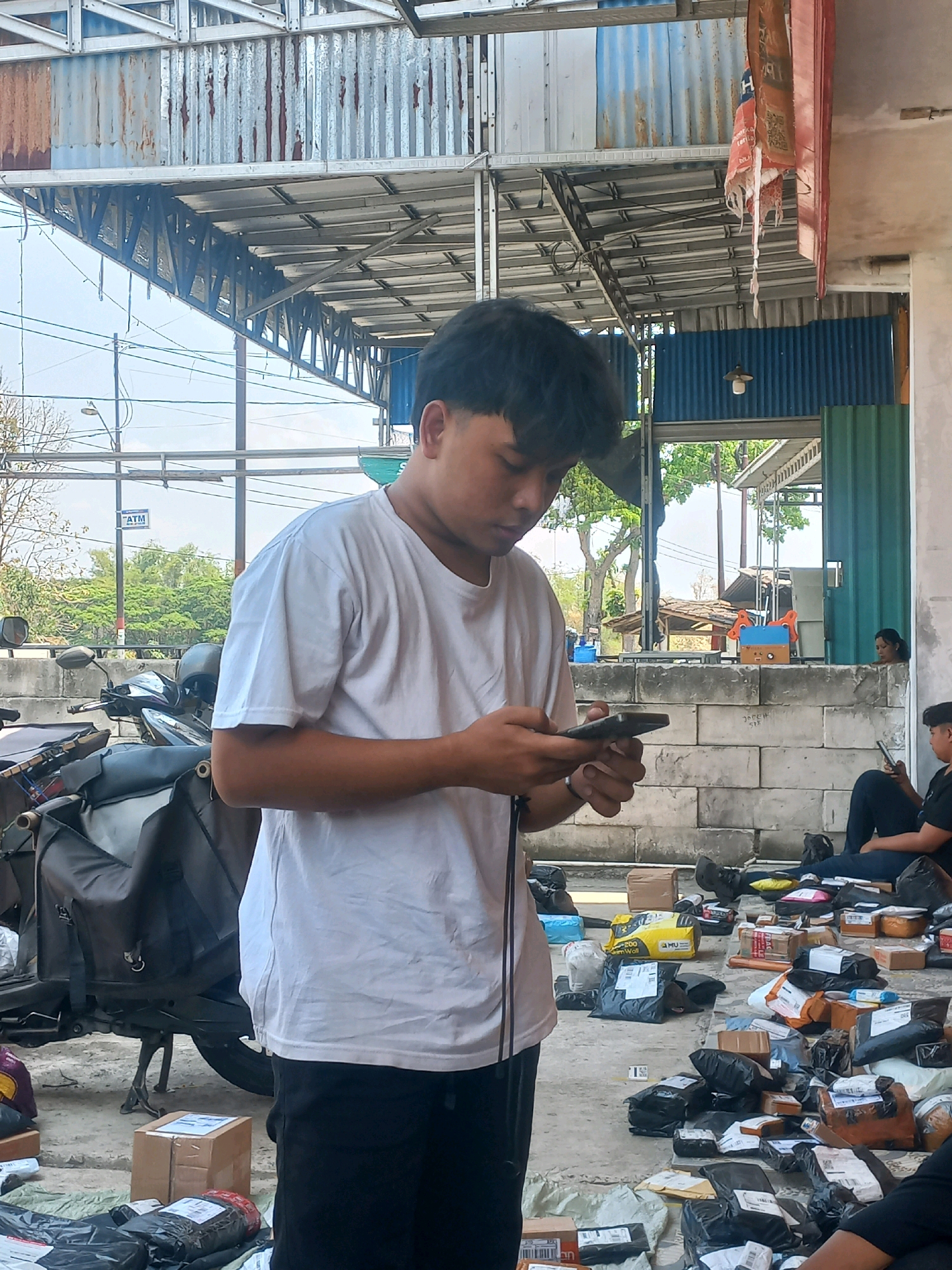 In Baturetno, Indonesia, a young man stands outside, looking down at his phone. He is wearing a white t-shirt and dark pants. The environment is an open area, likely a warehouse or delivery hub, with a corrugated metal roof supported by metal beams overhead. A motorcycle is parked to his left, with a large black bag attached. Numerous packages of various sizes are scattered on the ground around him, suggesting a logistics or shipping operation. In the background, a wall constructed of cinder blocks separates the area from a more enclosed space. Other individuals can be seen in the background, with one person sitting on the ground also looking at a phone, and another partially visible sitting near the wall. The overall scene depicts a busy, outdoor workspace related to package handling and distribution.