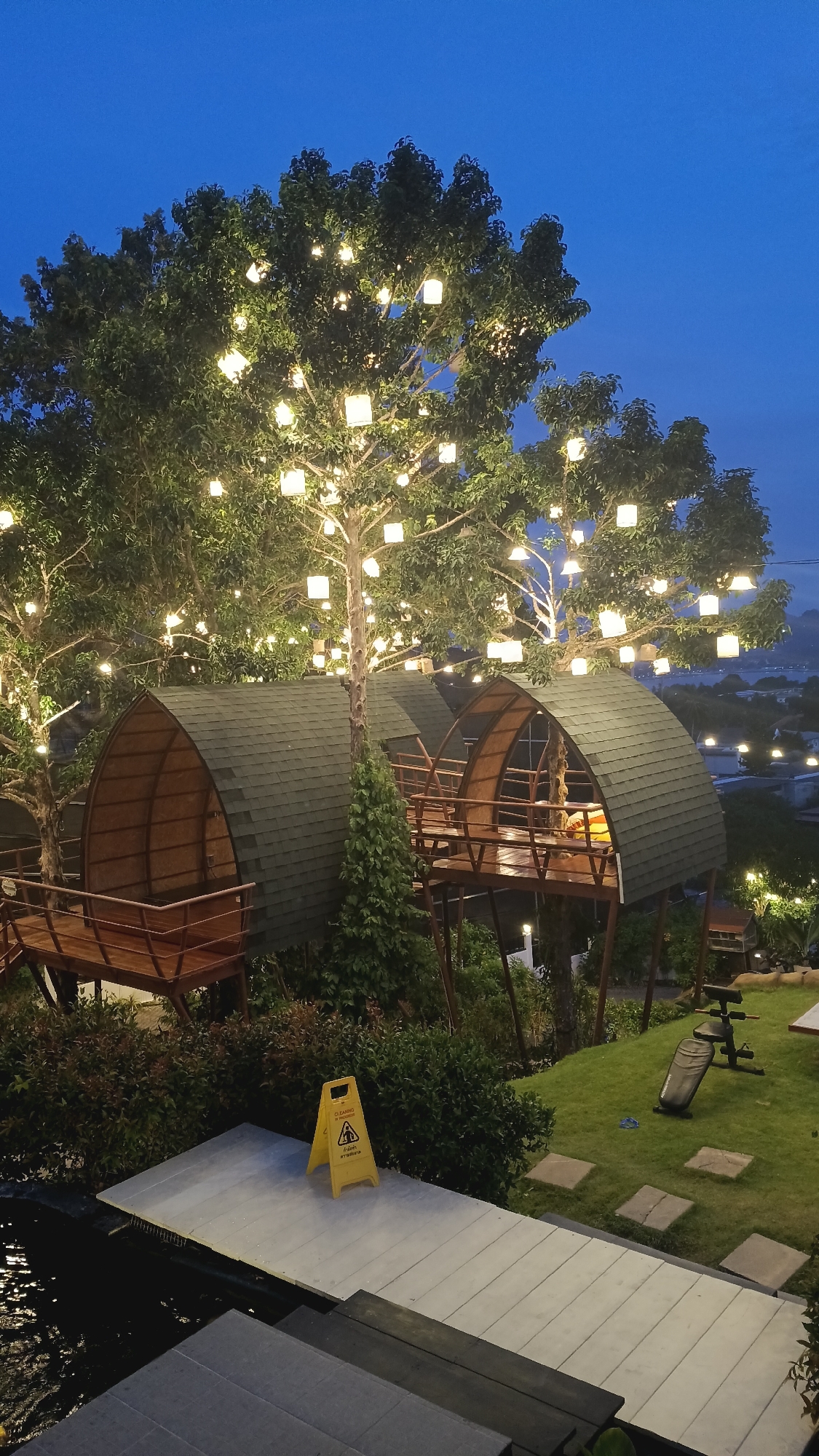 In the city of Ko Samui, Thailand, a resort or park area is depicted at dusk. Two distinctive, dome-shaped wooden structures, reminiscent of treehouses or pods, are elevated on stilts and feature sloped, dark green shingled roofs. These structures have wooden decks and railings, with visible interiors. They are nestled amongst lush green trees adorned with numerous square, white lanterns that emit a warm glow. The background reveals a cityscape with buildings and lights, suggesting an urban or semi-urban setting, and a body of water. In the foreground, a pathway made of light grey, rectangular planks leads towards a dark body of water. A yellow, triangular "wet floor" sign stands on the pathway. A grassy area to the right features a weight bench and stepping stones. The overall ambiance is tranquil and illuminated by the ambient twilight and the decorative lanterns.