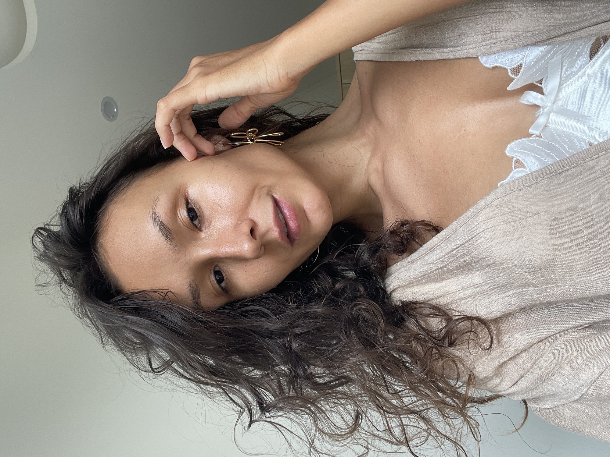The image shows a close-up of a woman with dark, curly hair and fair skin. She is wearing a beige cardigan over a white lace bralette. She has a gold bow-shaped earring in her right ear and is touching her face with her right hand. The background is a plain, light-colored wall with a circular light fixture visible on the ceiling. The image is lit from the front, highlighting her features and the textures of her clothing and hair. There is no indication of the location being in Mũi Né, Vietnam.