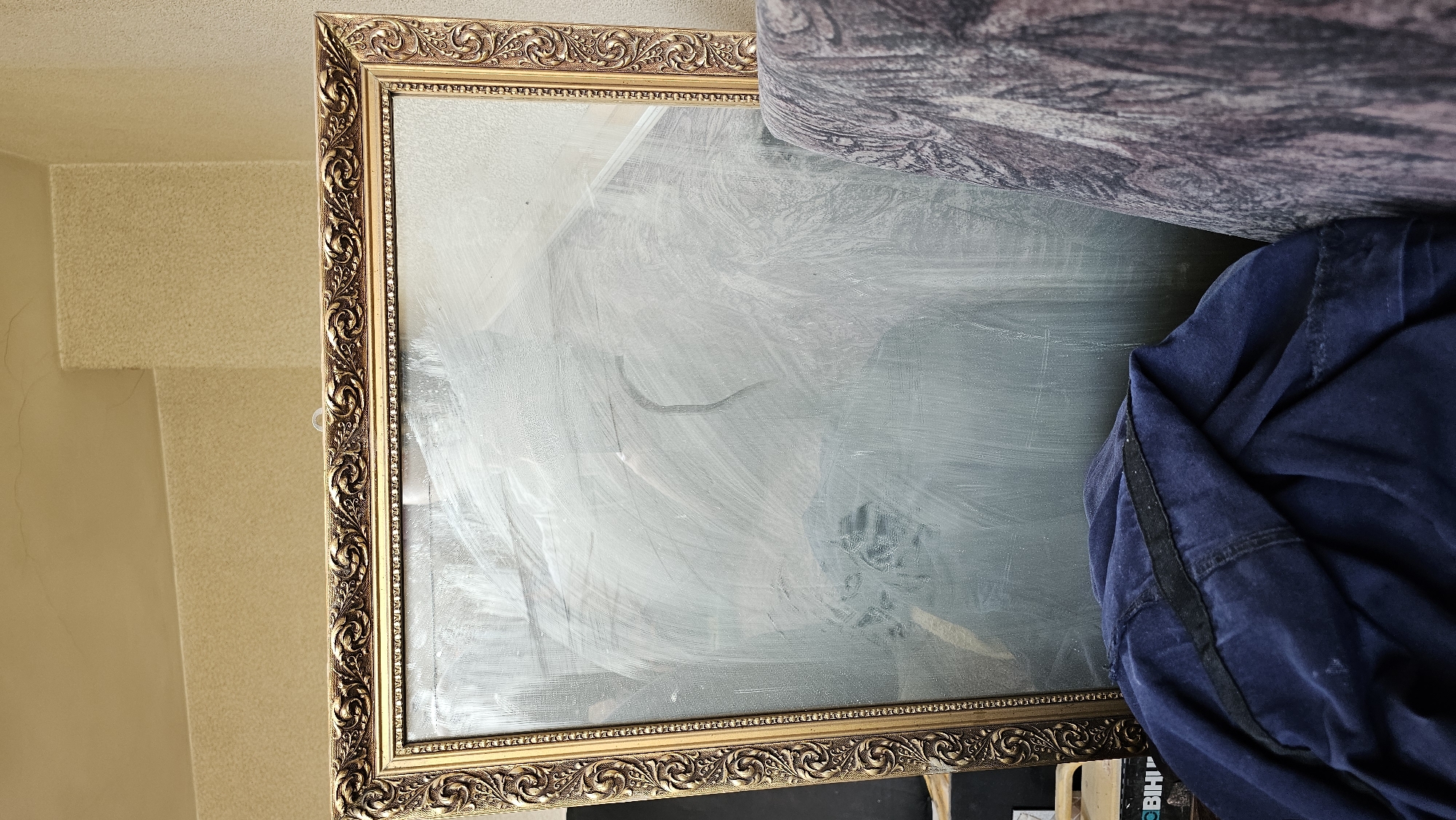 The image depicts a large, ornate gold-framed mirror leaning against a textured wall, possibly in a residential setting in Sofia, Bulgaria. The mirror's surface is smudged with white streaks, suggesting it has recently been cleaned or painted. To the left of the mirror, a dark blue garment is draped over an unseen object, partially obscuring the lower portion of the frame and the mirror's reflection. To the right of the mirror, a portion of a patterned armchair or sofa is visible, with a dark, possibly brown and grey, fabric featuring a textured design. The wall behind the mirror is a light beige color with a subtle textured finish. The overall impression is of a room undergoing some form of transition or storage, with items temporarily placed.