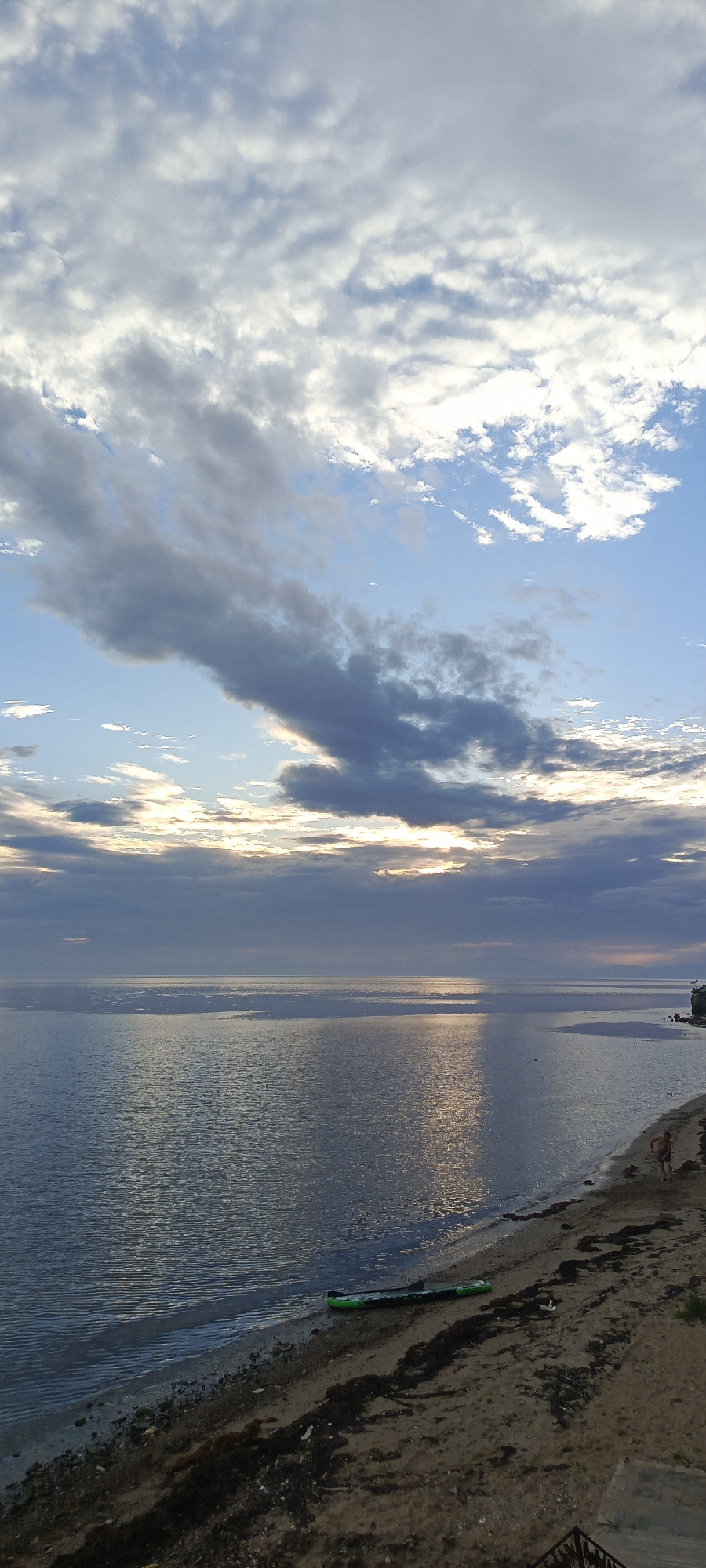 The image depicts a broad expanse of calm water, likely the Sea of Japan, meeting a sandy and rocky shoreline. The sky is partly cloudy with dramatic formations of grey and white clouds interspersed with patches of blue sky, suggesting either sunrise or sunset. Sunlight filters through the clouds, creating a shimmering reflection on the water's surface.
On the shore, a green inflatable kayak or paddleboard rests on the sand. Further up the beach, a person, appearing to be a male wearing dark shorts, stands facing the water. Another figure, also appearing to be a male, is partially visible further down the beach, near a rocky outcrop on the right side of the frame. The shoreline is sparsely populated with seaweed and debris. The overall impression is one of a tranquil coastal scene. The location is identified as Bolshoy Kamen, Russia.