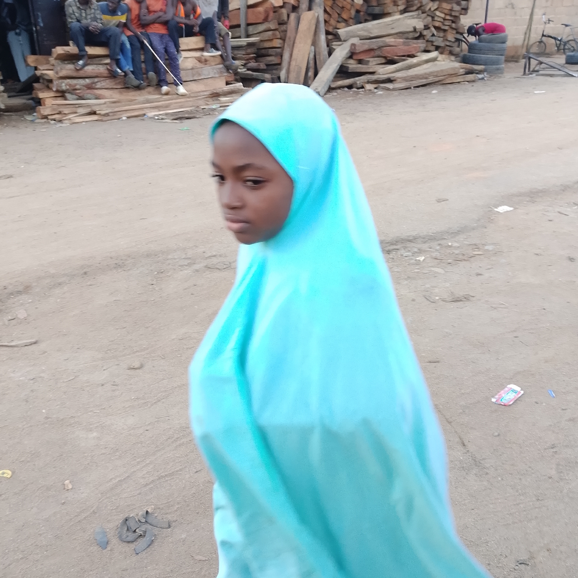The image is taken from a medium-low angle, showing a young girl in a bright blue hijab in the foreground, looking to her left and slightly downwards. Her face is partially visible and in focus. Behind her, the background is blurred but shows an outdoor scene in what appears to be a rural or informal urban setting. A pile of logs is visible on the left, and several people are seated on these logs. Further in the background, a stack of tires is present, with a person in a red garment perched on top. A bicycle and a ramp are also visible in the distance. The ground is dirt and appears unpaved. There are some scattered debris and objects on the ground in the middle and right portions of the image. The lighting suggests it is daytime, possibly overcast or late afternoon.