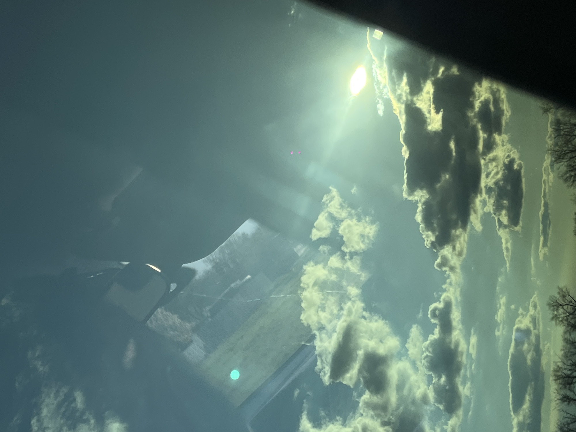 The image captures a bright, cloudy sky scene from the interior of a vehicle, likely a car, looking through its windshield. The sky is a mix of blue-grey tones with prominent, textured clouds, particularly dense on the right side of the frame. The sun, a brilliant white light source, is visible in the upper-right quadrant, creating a strong lens flare effect across the sky. Two small, distinct magenta-purple dots appear in the upper-central sky, positioned between the sun and the main cloud mass.
Bare tree branches are visible along the far right edge of the image, suggesting a colder season. The interior of the vehicle is partially reflected or visible through the windshield. The upper-left portion of the frame shows a dark, blurry area, indicative of the car's interior. A large, rectangular reflection, likely from an infotainment screen or a mounted mobile device, dominates the lower-left to central part of the view. Within this reflection, a bright, circular teal light is clearly visible. Faint details of a landscape, possibly rural with distant fields and structures, can be seen through the windshield, beyond the reflections. The scene depicts a sunny day with significant cloud cover, observed while in transit or stationary in a vehicle. No visible text is present.