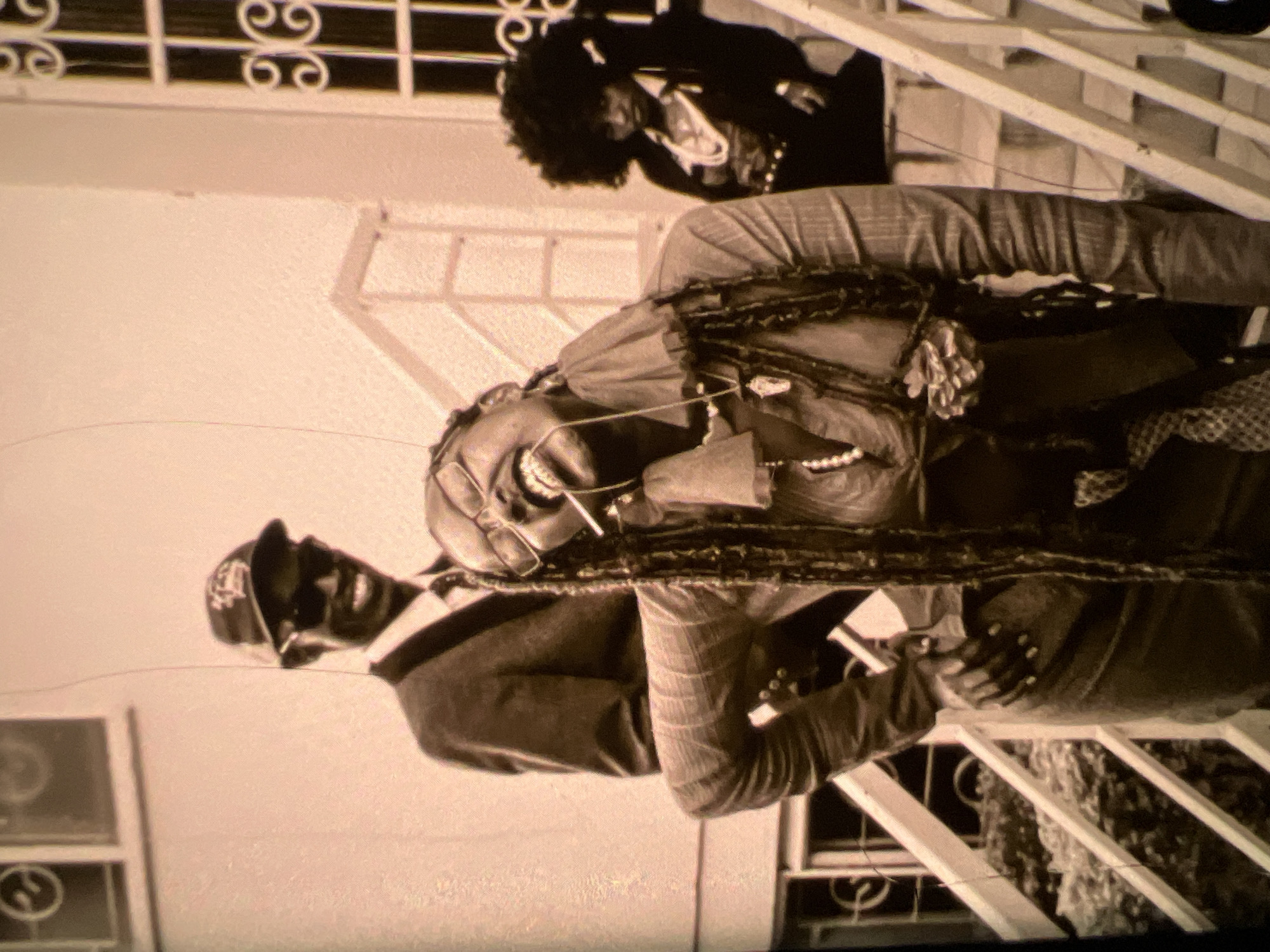 This is a black and white, grainy, medium shot of three individuals on a staircase. The central figure, a woman with long braids and glasses, is laughing with her head tilted back. She is wearing a button-up shirt with a ruffled collar and a necklace. She is holding a cigarette in her mouth and her left hand is on her hip. Behind her and to the left, a man in a suit and baseball cap is smiling. To the right and further back, a woman with an afro is looking towards the camera. The staircase is made of wood and painted white, with a railing. The background appears to be a building with windows and decorative ironwork. The overall setting suggests an outdoor gathering in a residential or public space, possibly in Lagos, Nigeria, given the individuals' appearances.