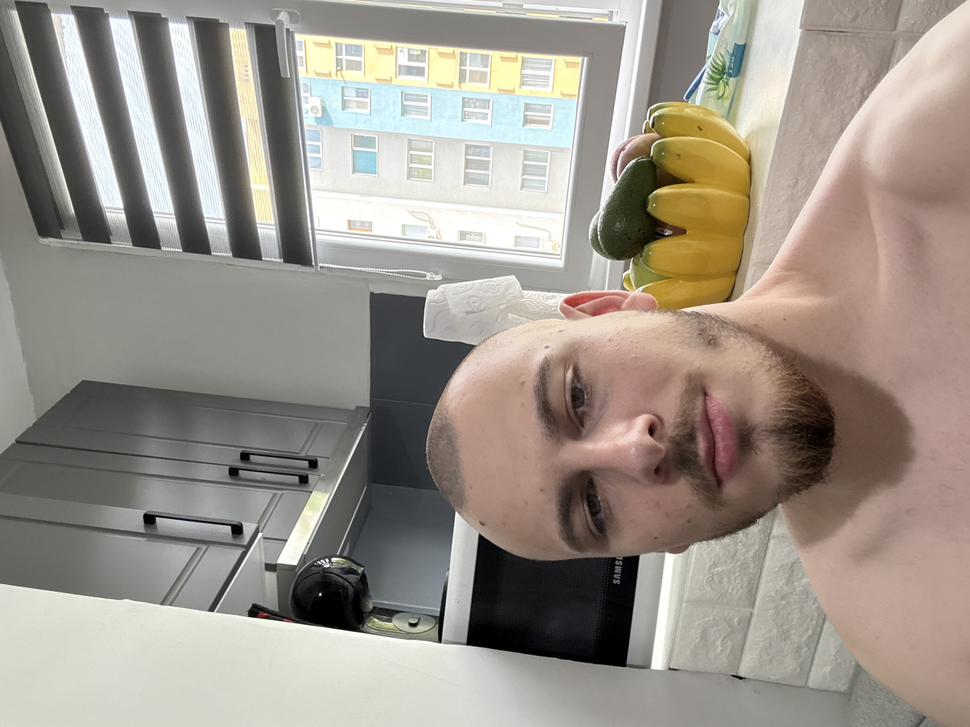 A shirtless man with a short haircut and a beard is in a kitchen. Behind him are kitchen cabinets, a microwave oven, a window with blinds, and a building with a blue and yellow facade visible through the window. The building visible is likely in Stara Zagora, Bulgaria, given the user's request, although without further information this cannot be confirmed. There is a banana-shaped bowl with fruit on the counter.