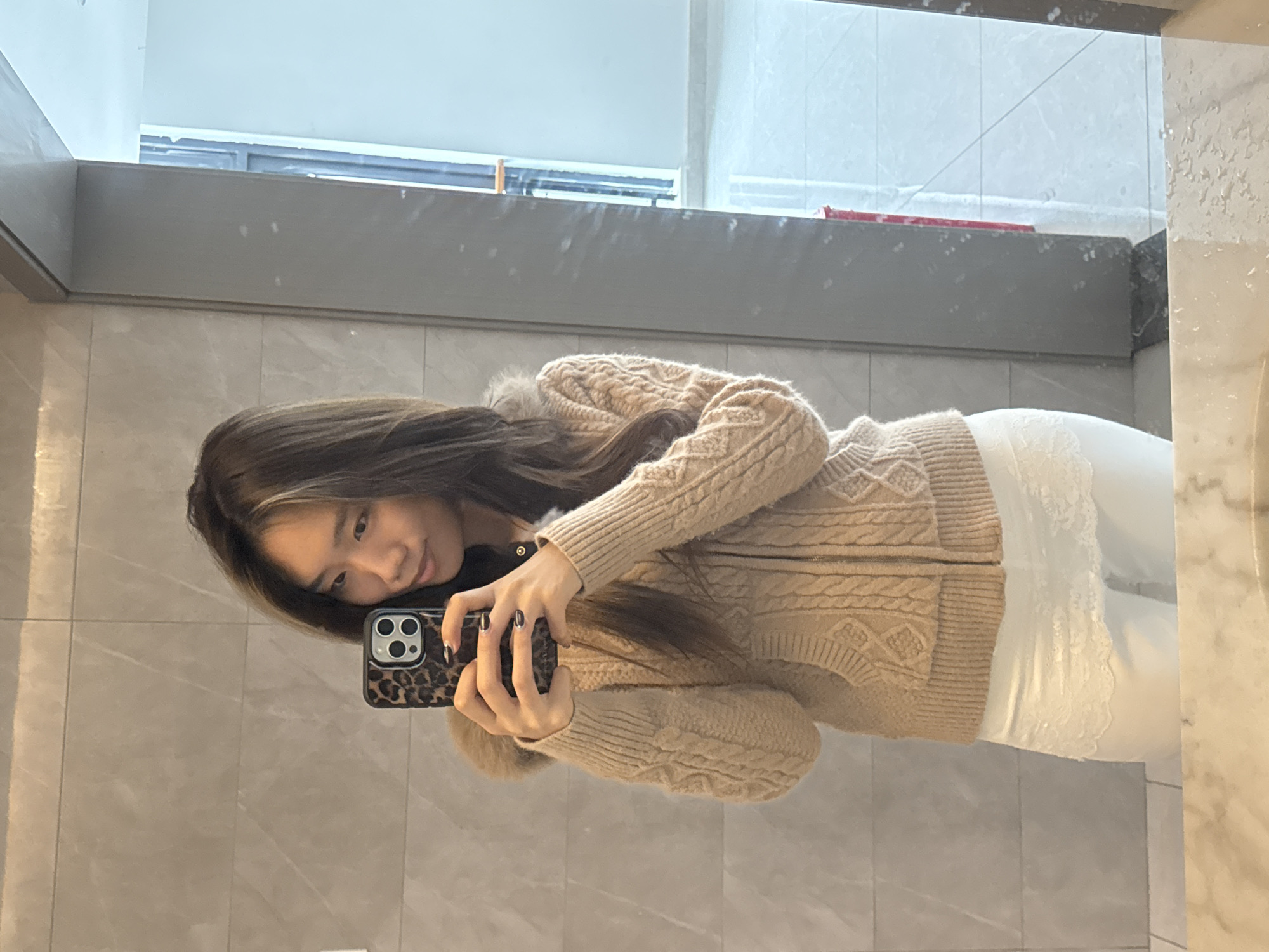 A young Asian woman is taking a selfie in what appears to be a bathroom. She is wearing a beige knitted cardigan and white pants with lace detailing. She is holding a smartphone with a leopard print case. The bathroom has gray tiles and a large mirror, with a window visible in the upper background. The location appears to be the Nan'an District in China.