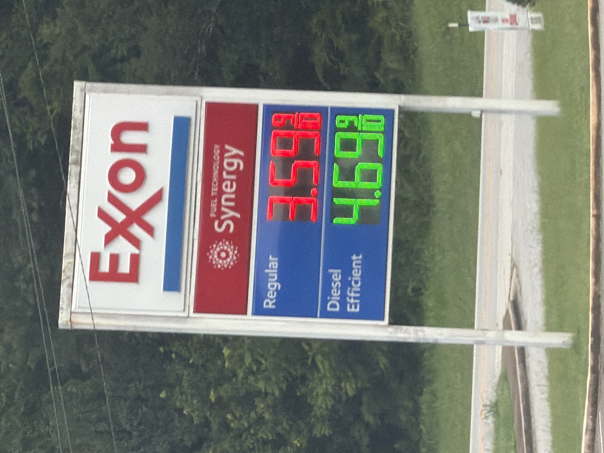 An outdoor gas station sign is visible in Wildwood, United States. The sign, mounted on a white pole, displays the Exxon logo at the top in red lettering. Below the logo, "FUEL TECHNOLOGY Synergy" is written on a red background. The sign then shows prices for different types of fuel. "Regular" fuel is priced at 3.59 9/10 dollars per gallon, displayed in red digital numbers. "Diesel Efficient" fuel is priced at 4.69 9/10 dollars per gallon, displayed in green digital numbers. The background consists of lush green trees and foliage. The sign is positioned in front of a grassy area with a road visible in the lower portion of the image.