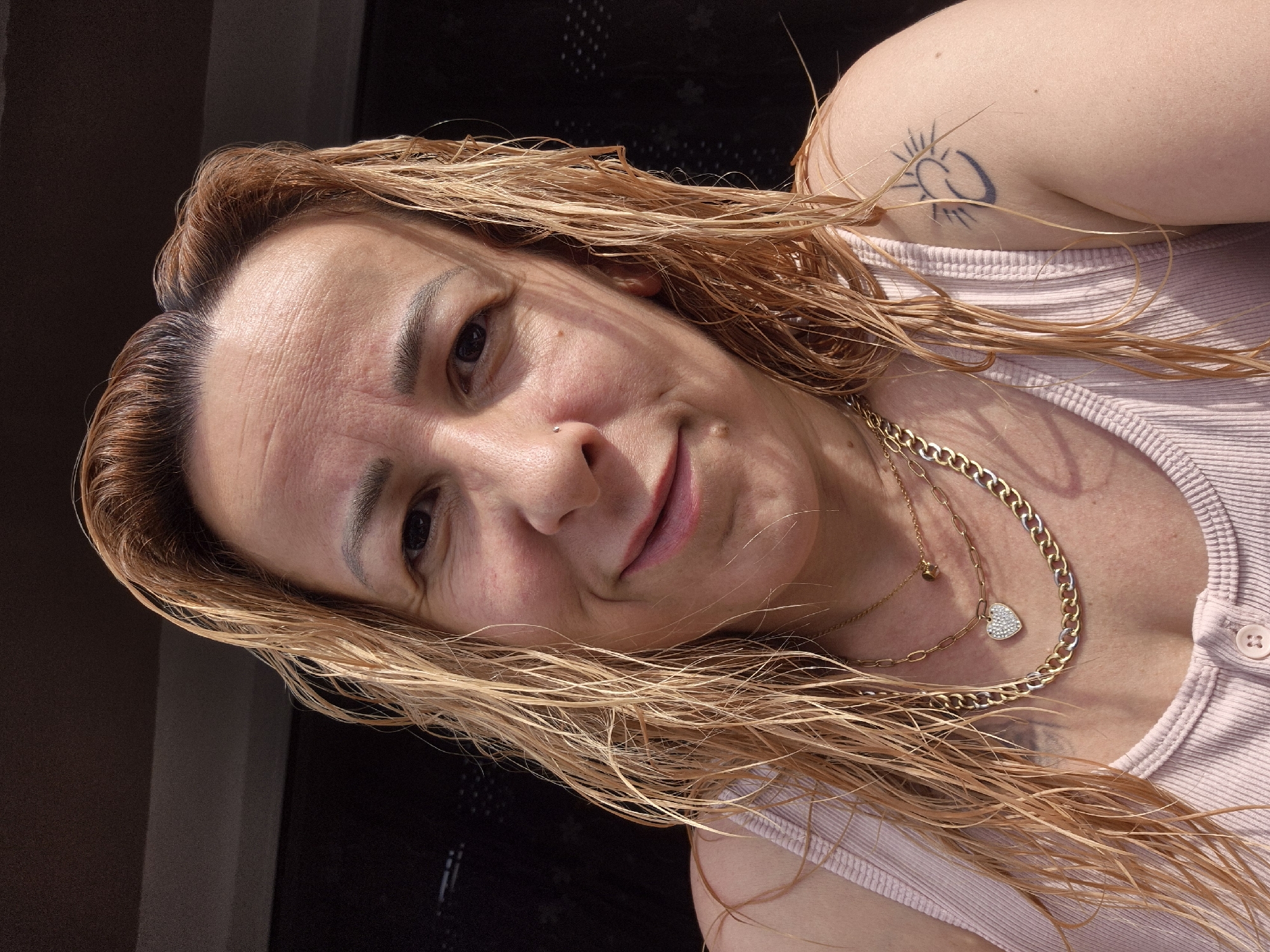 An outdoor, medium-shot, close-up selfie shows a light-skinned woman with blonde, wet hair. She is looking directly at the camera, smiling, and has a nose ring and several necklaces. Her left shoulder is visible, and a tattoo of a sun and crescent moon is on her upper arm. She is wearing a light pink tank top. The background is dark and out of focus.
