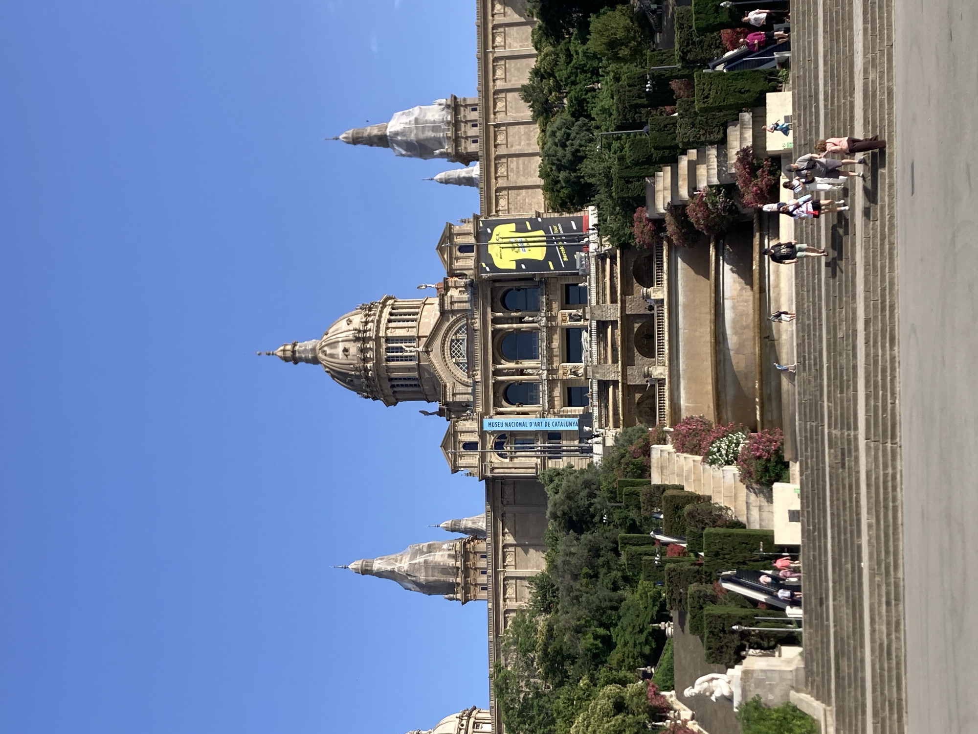 This is a frontal, eye-level, full shot of the Palau Nacional in Barcelona, Spain, which houses the National Art Museum of Catalonia. The iconic building is under a clear blue sky. In the foreground, a wide, multi-tiered stone staircase descends from the palace. People are walking up and down the stairs. Some are in small groups, others are alone. To the left of the stairs, there is a sculpted statue of a woman. Behind the statue, an escalator ascends between two flights of stairs. The stairs are flanked by manicured hedges and flowering bushes, with a tiered fountain visible behind them. Two large banners hang from the palace facade; one is a yellow and black advertisement, and the other is a blue banner with white text. The palace itself is grand, with a large central dome and smaller domes and spires on either side, all made of stone and adorned with architectural details.